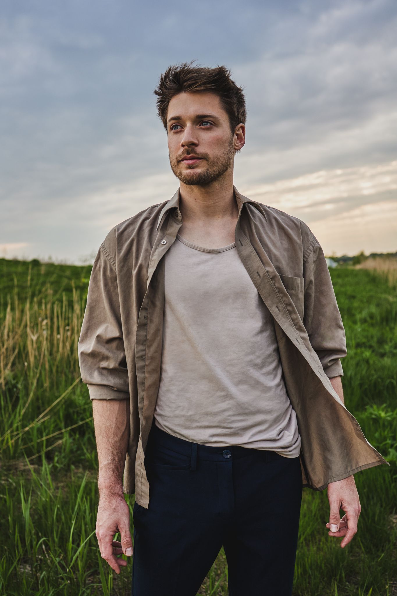 A person in a beige jacket and white t-shirt stands in a field of tall grass during an overcast evening.