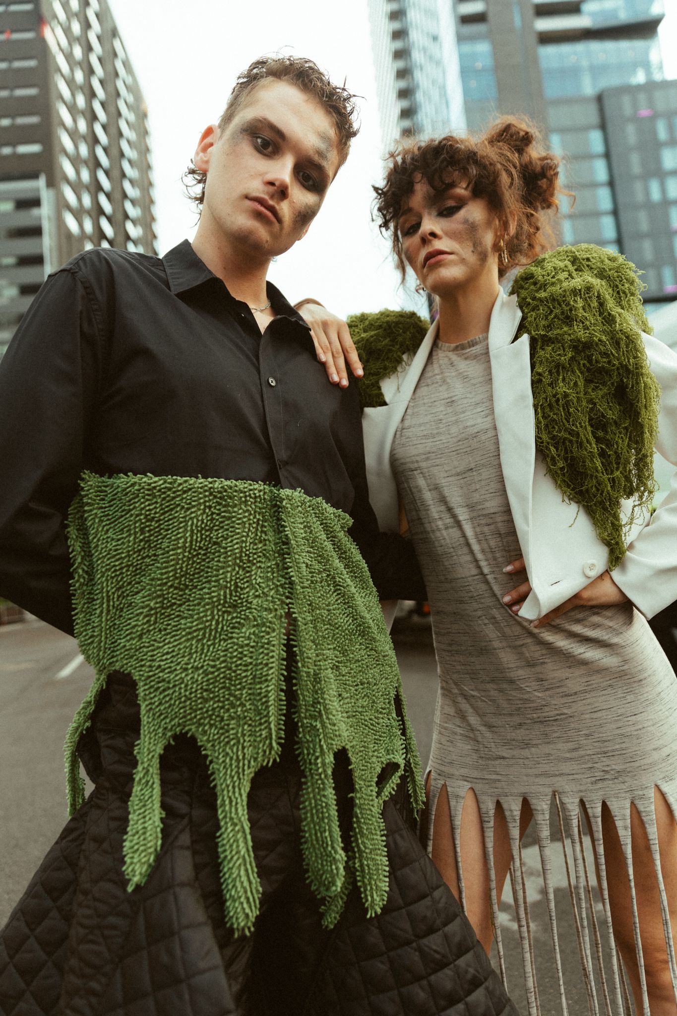 Two models pose in avant-garde fashion featuring green moss-like textures and urban streetwear against city backdrop.