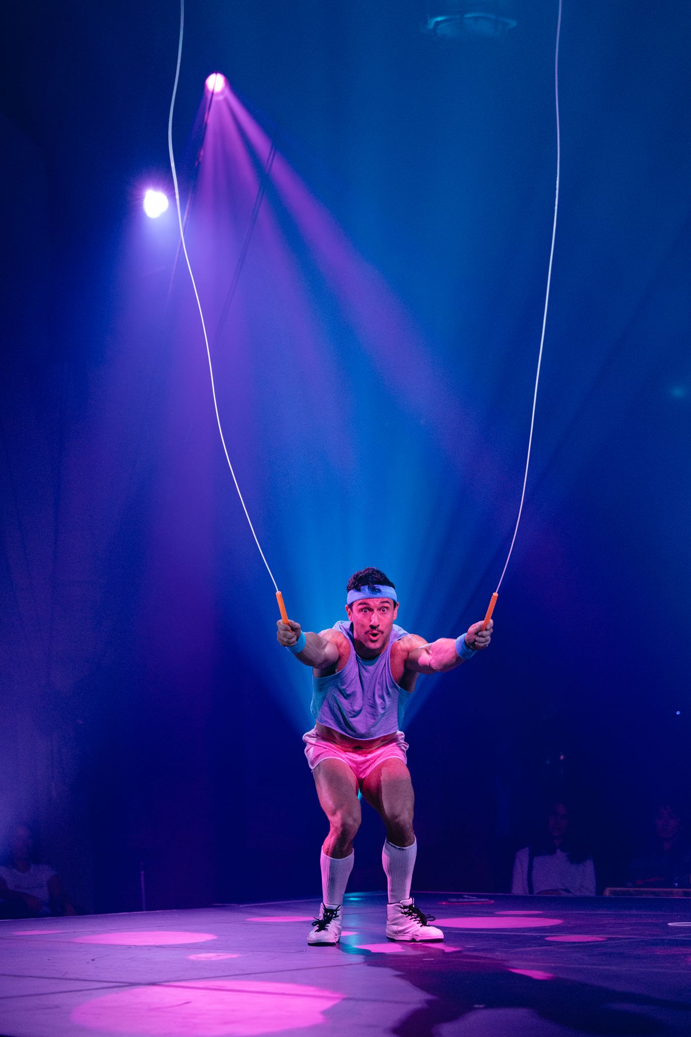 Performer in casual athletic wear swings on illuminated stage ropes under dramatic purple and blue spotlights.