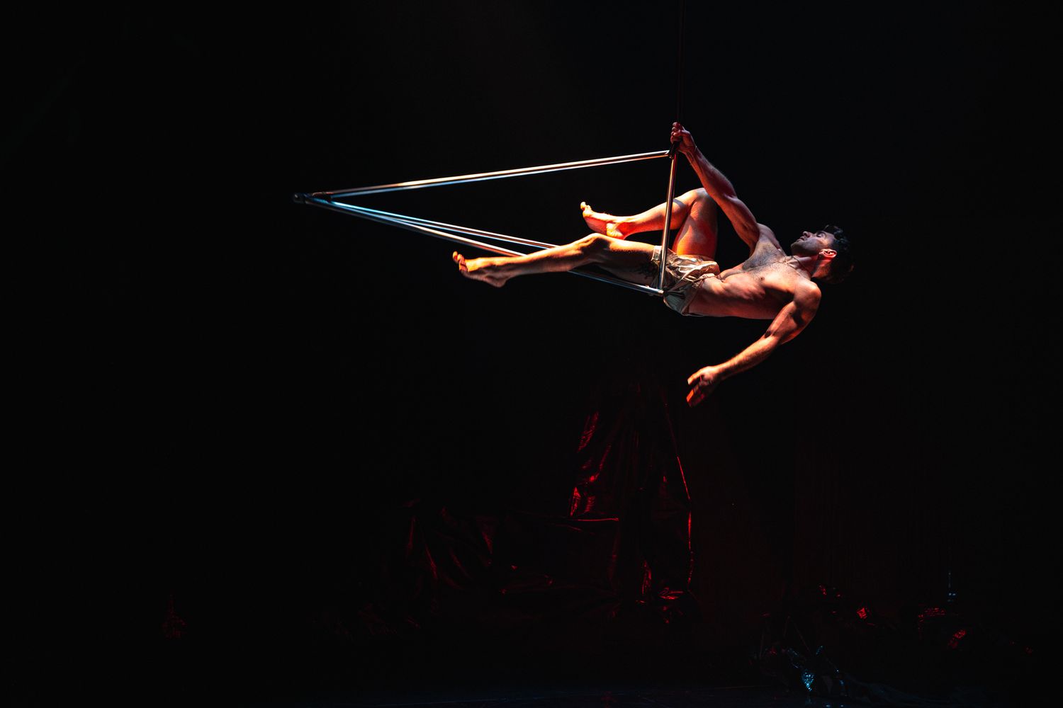 An aerial performer in a red costume gracefully suspended on metal apparatus against dark background.