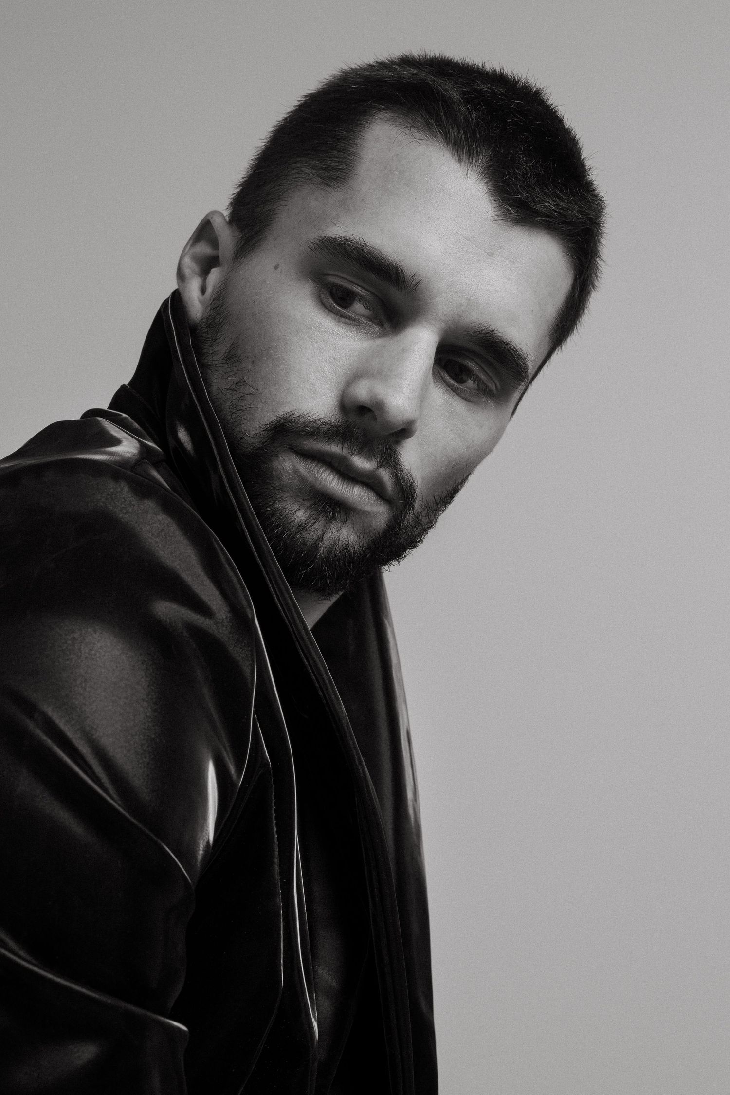 Black and white portrait of person wearing leather jacket looking over shoulder against plain background.