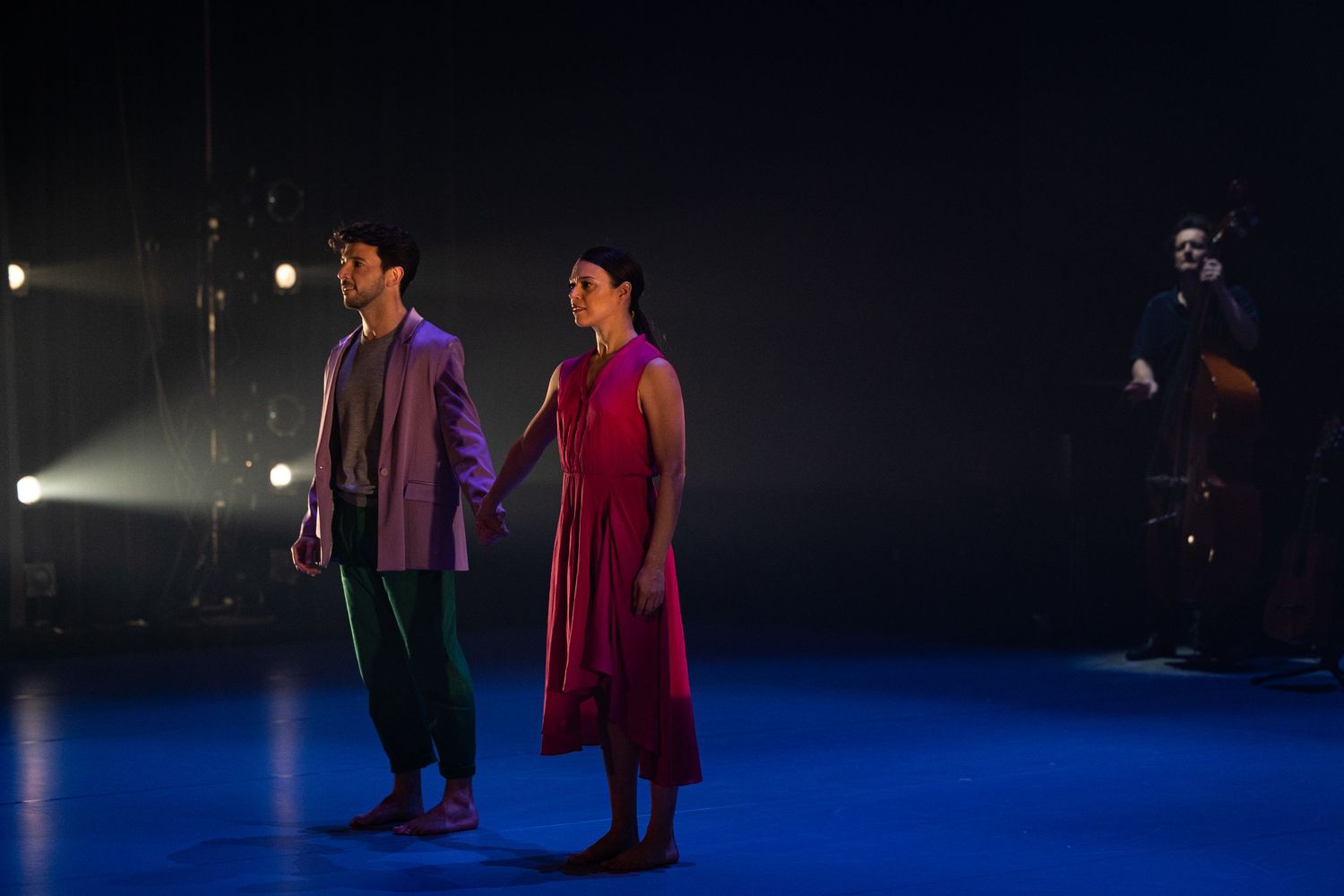 Performers in red and green costumes move across a dimly lit stage with blue lighting.
