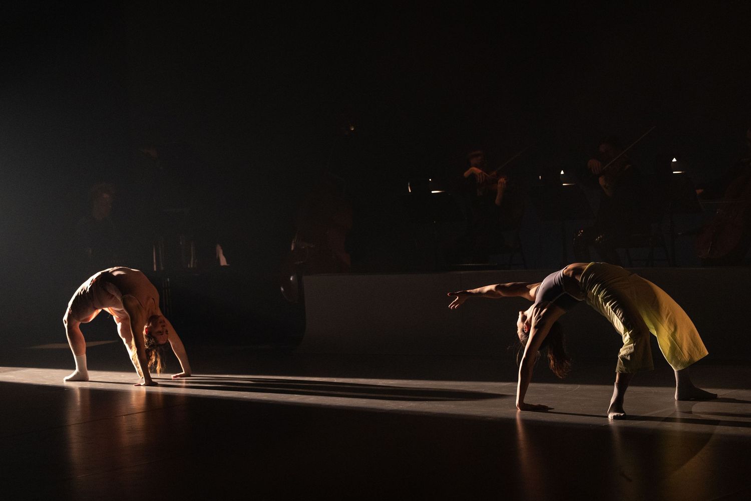 Two dancers perform a synchronized backbend move on a dimly lit stage illuminated by dramatic spotlights.