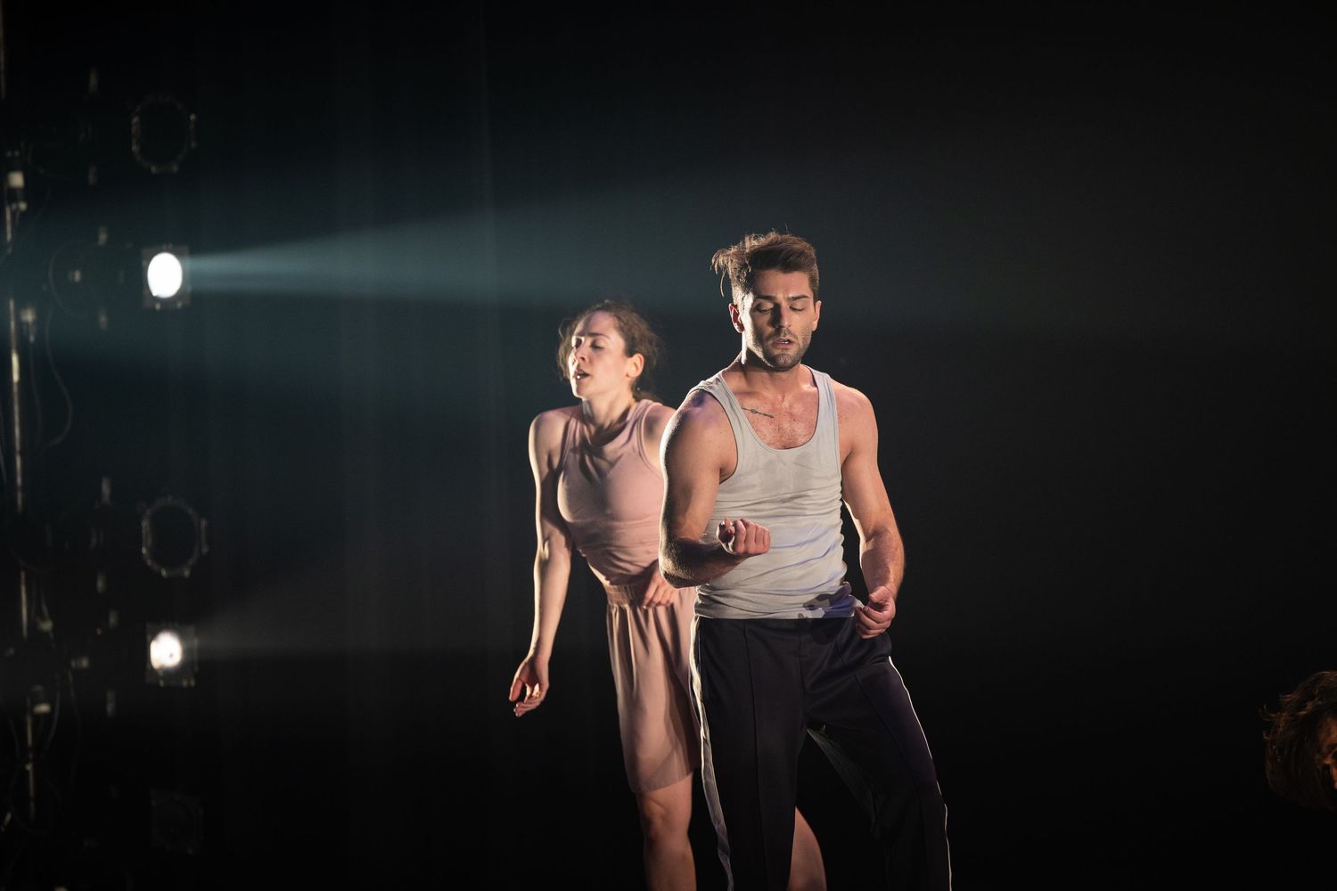 Two performers in dramatic lighting strike intense poses on a dark stage with theatrical spotlights beaming through fog.