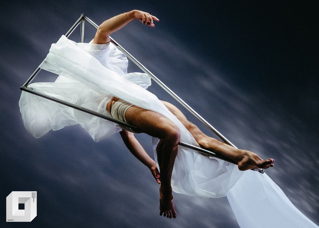 Artistic dancer in flowing white fabric creates dramatic pose against dark backdrop in striking studio photograph.
