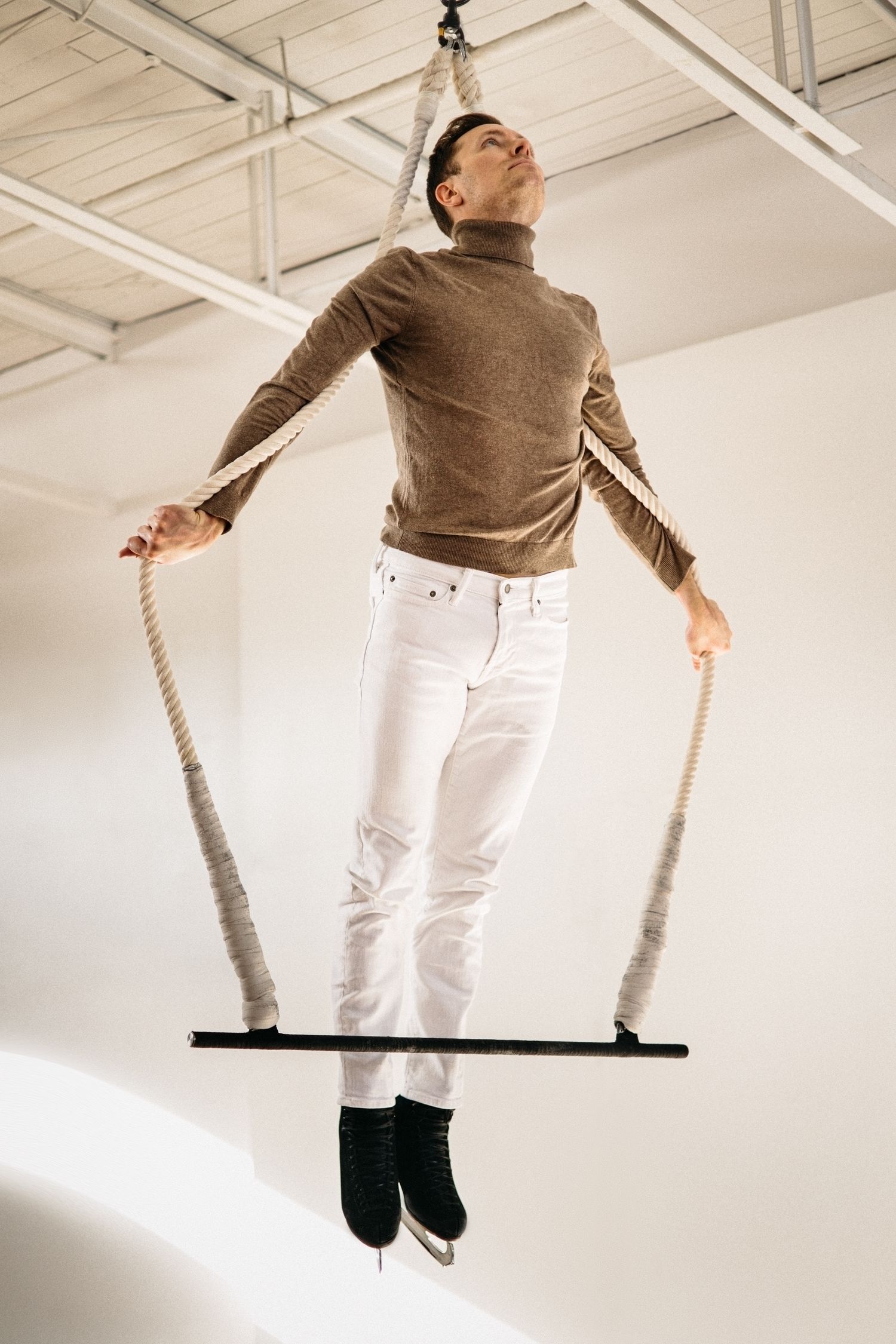 A person swinging upward on a suspended trapeze bar in sequence, wearing a brown sweater and white pants.