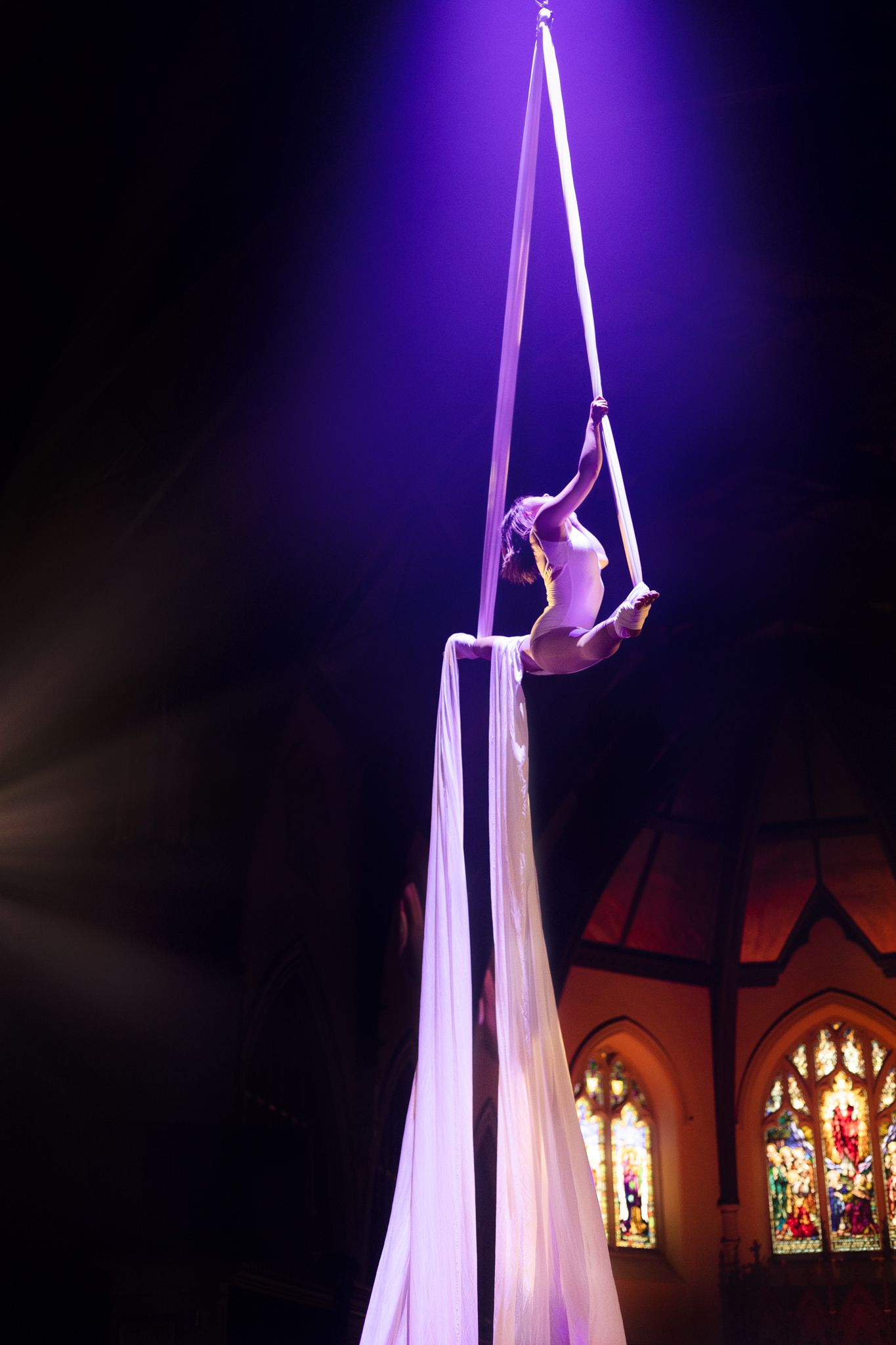 Aerial silk performer executes graceful pose on white fabric against purple lighting and stained glass windows.