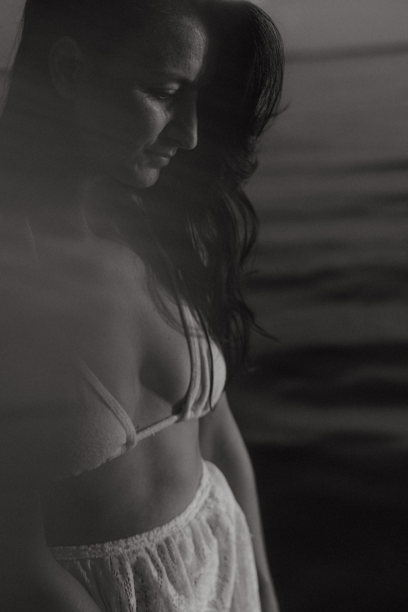 A moody black and white boudoir photo showing a silhouetted figure in delicate white lingerie against a dark background.