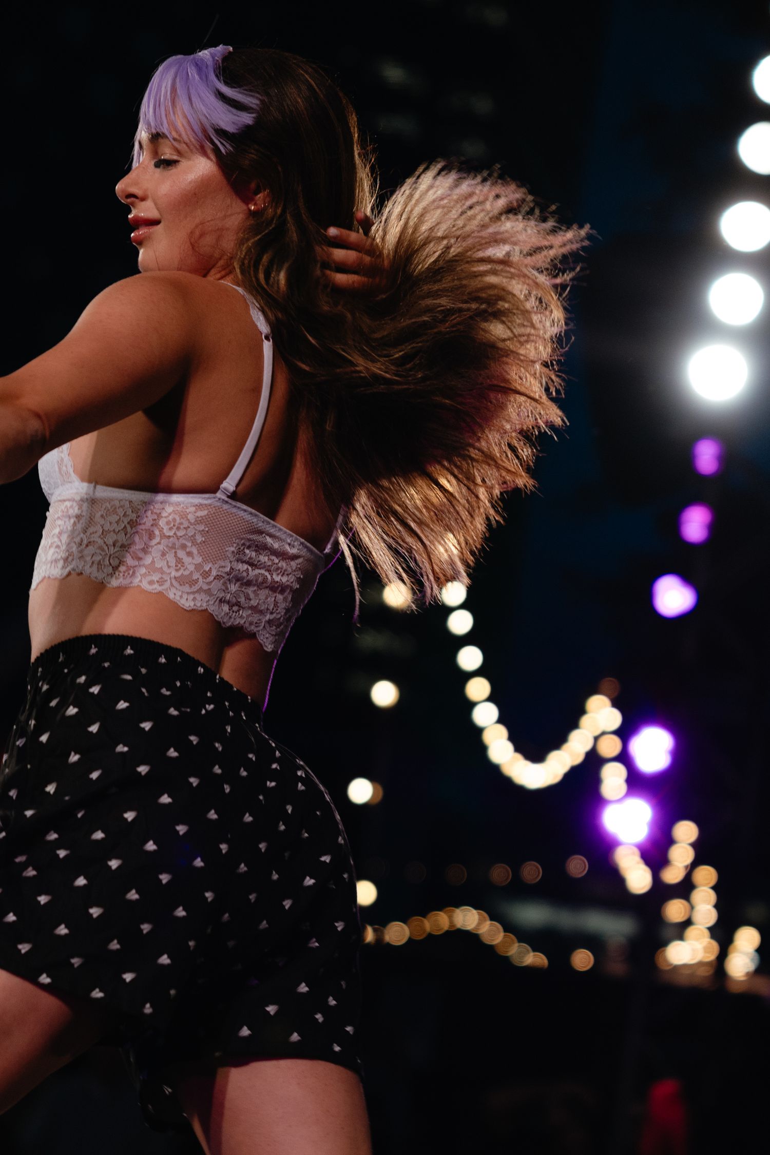 Performer in white lace top and polka dot skirt dances energetically on stage with hair flowing in motion.