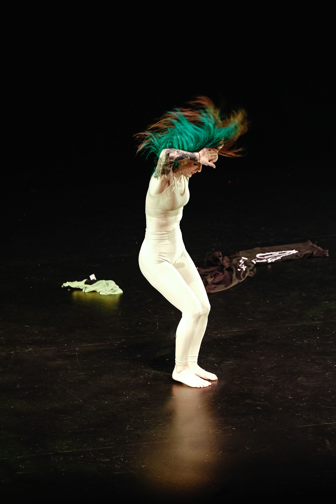 Dancer in white bodysuit performs dynamic movement with turquoise hair flowing on dark stage.