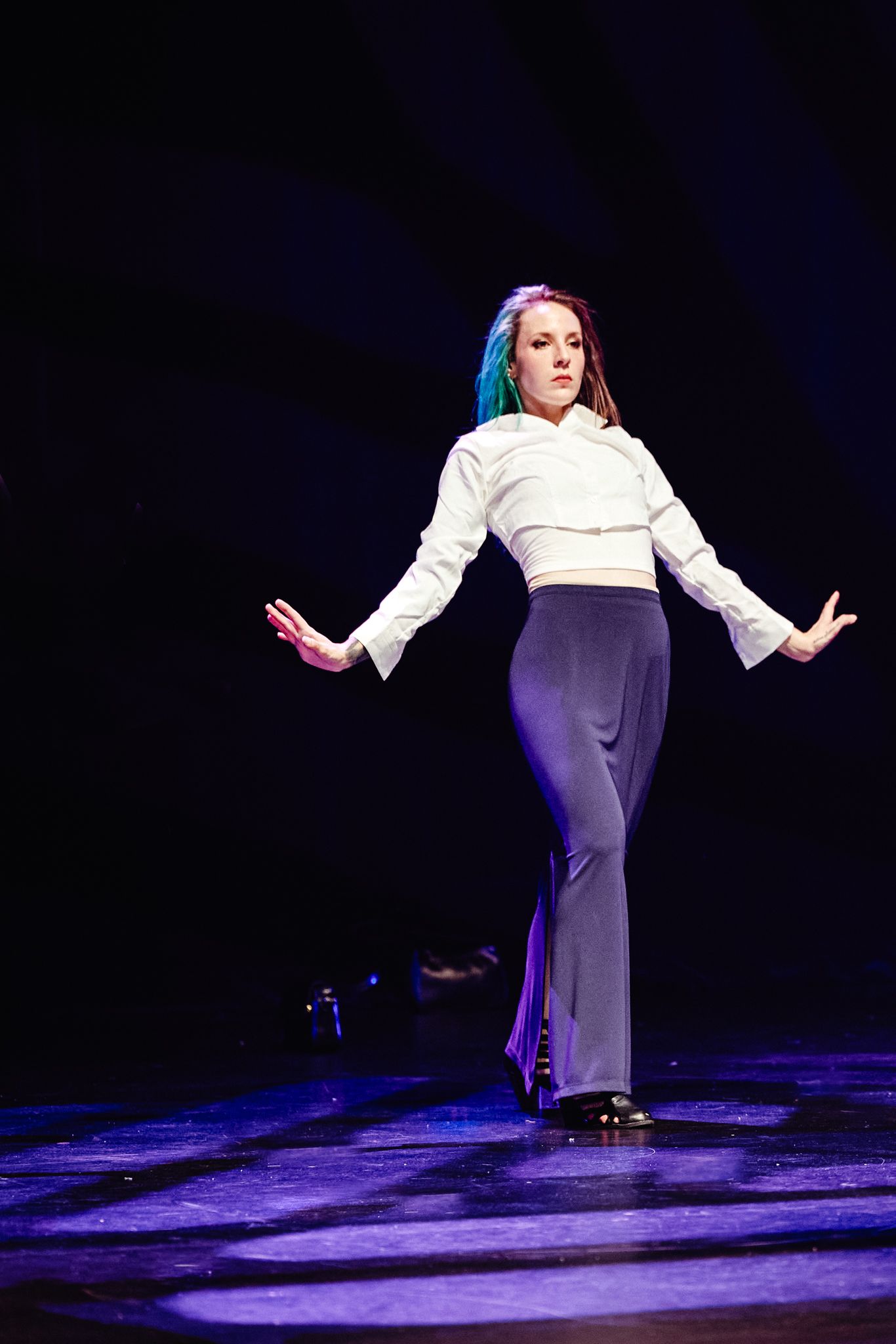 A performer in a white blouse and navy pants strikes an expressive pose under dramatic stage lighting.