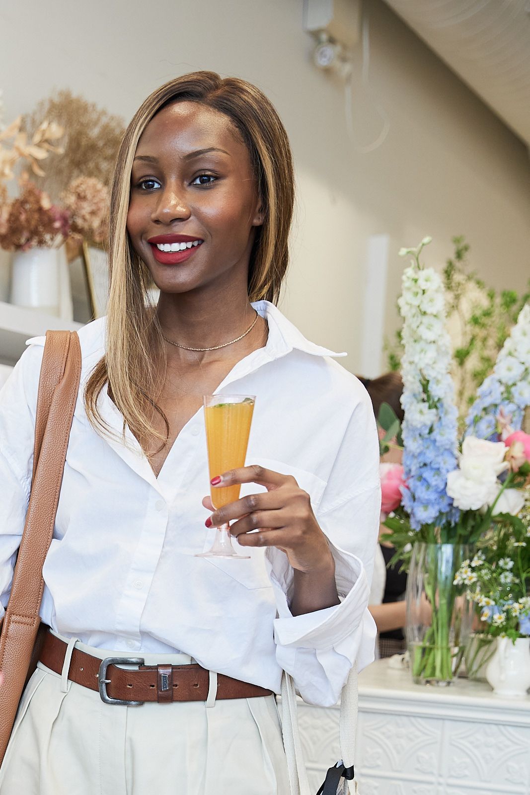 Woman in white button-down shirt holding a cocktail at an elegant event with floral decorations.