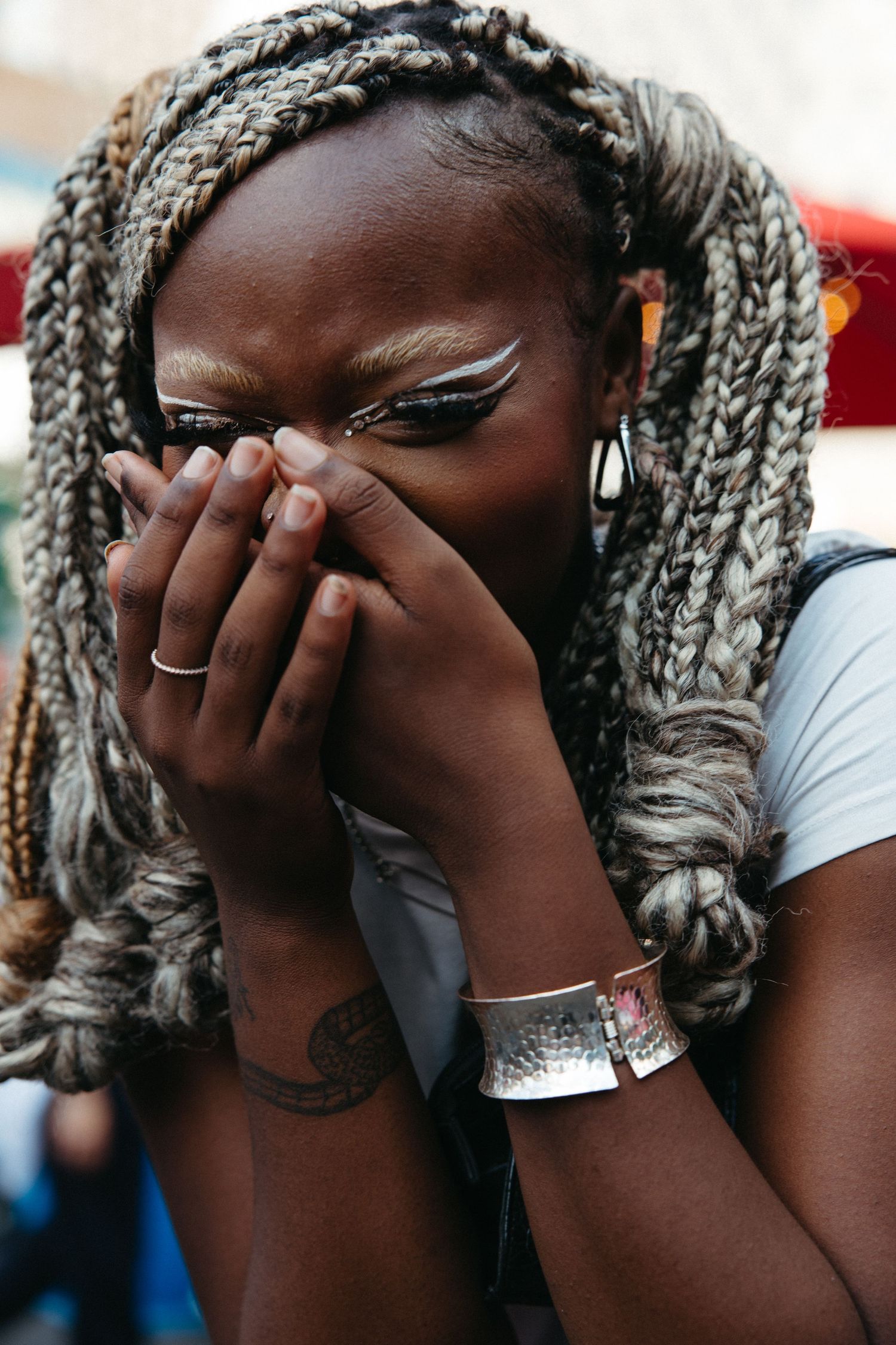 A joyful moment captured with stylish silver-gray braids, creative eye makeup, and hands covering a laughing expression.