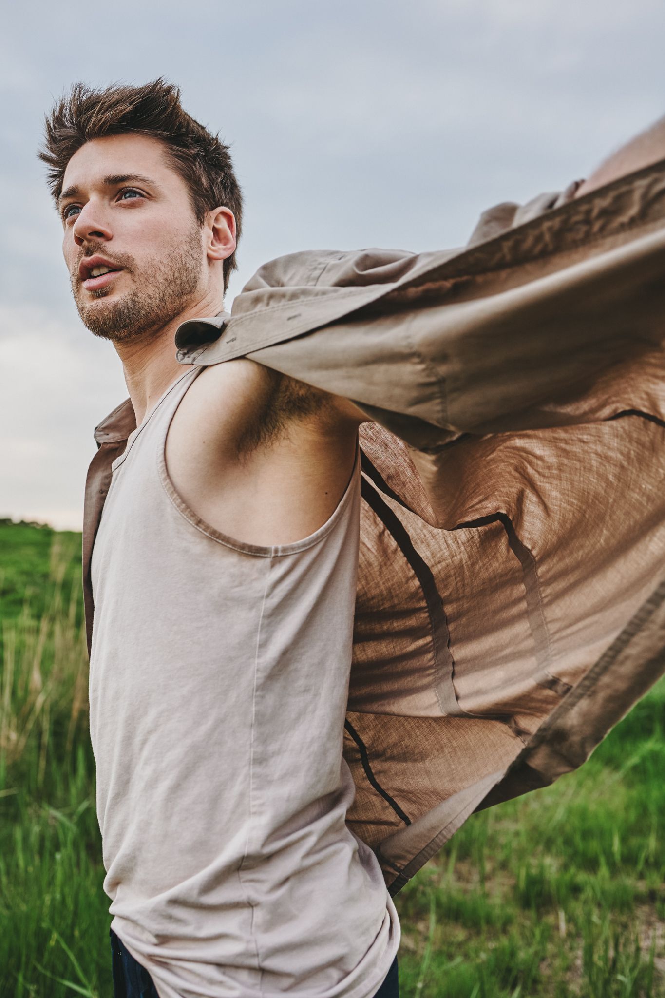 A person in a beige t-shirt and brown jacket spins joyfully in a grassy field against an overcast sky.