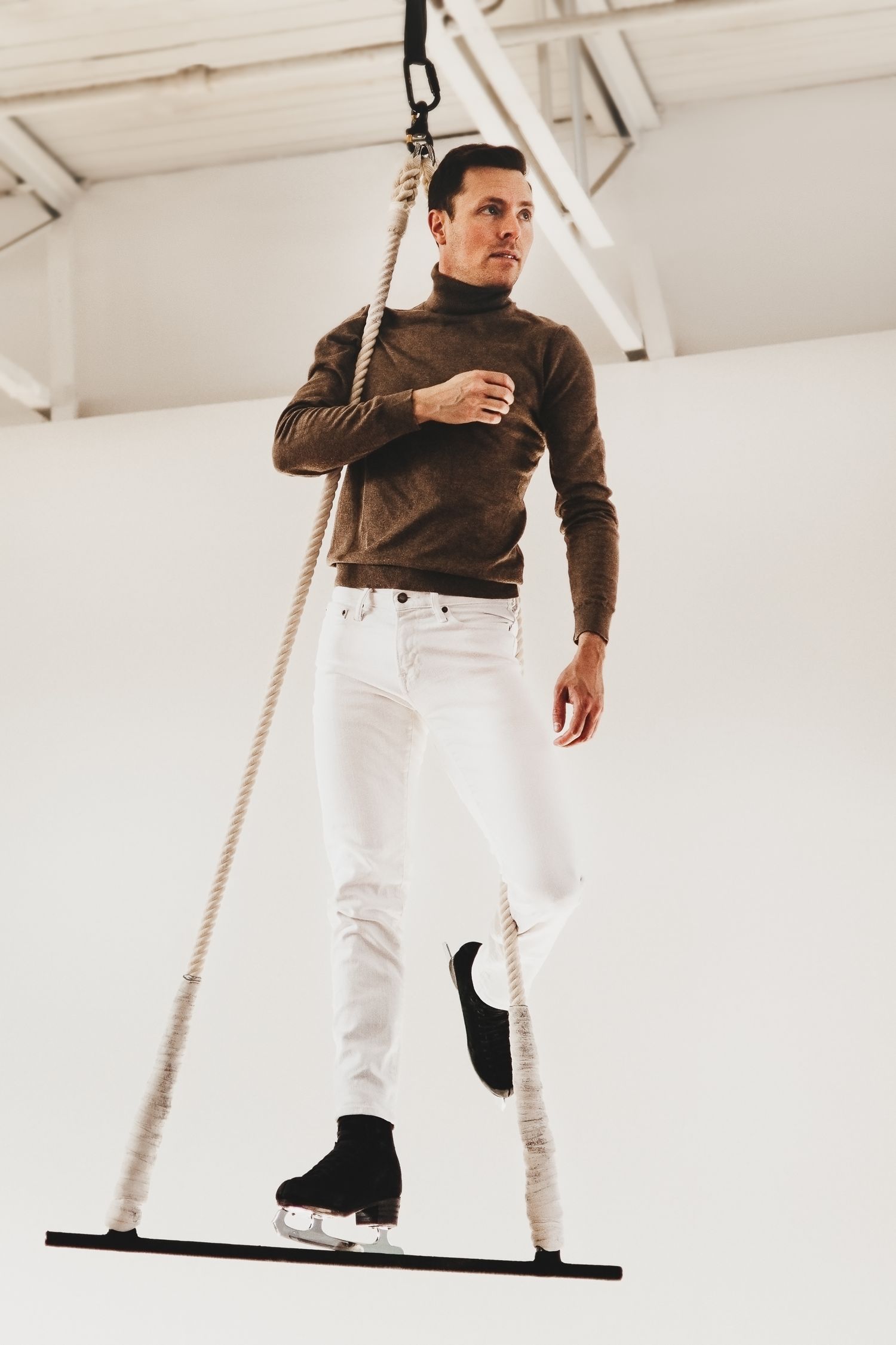 Stylish figure in brown turtleneck and white jeans balances on suspended platform in minimalist white studio space.
