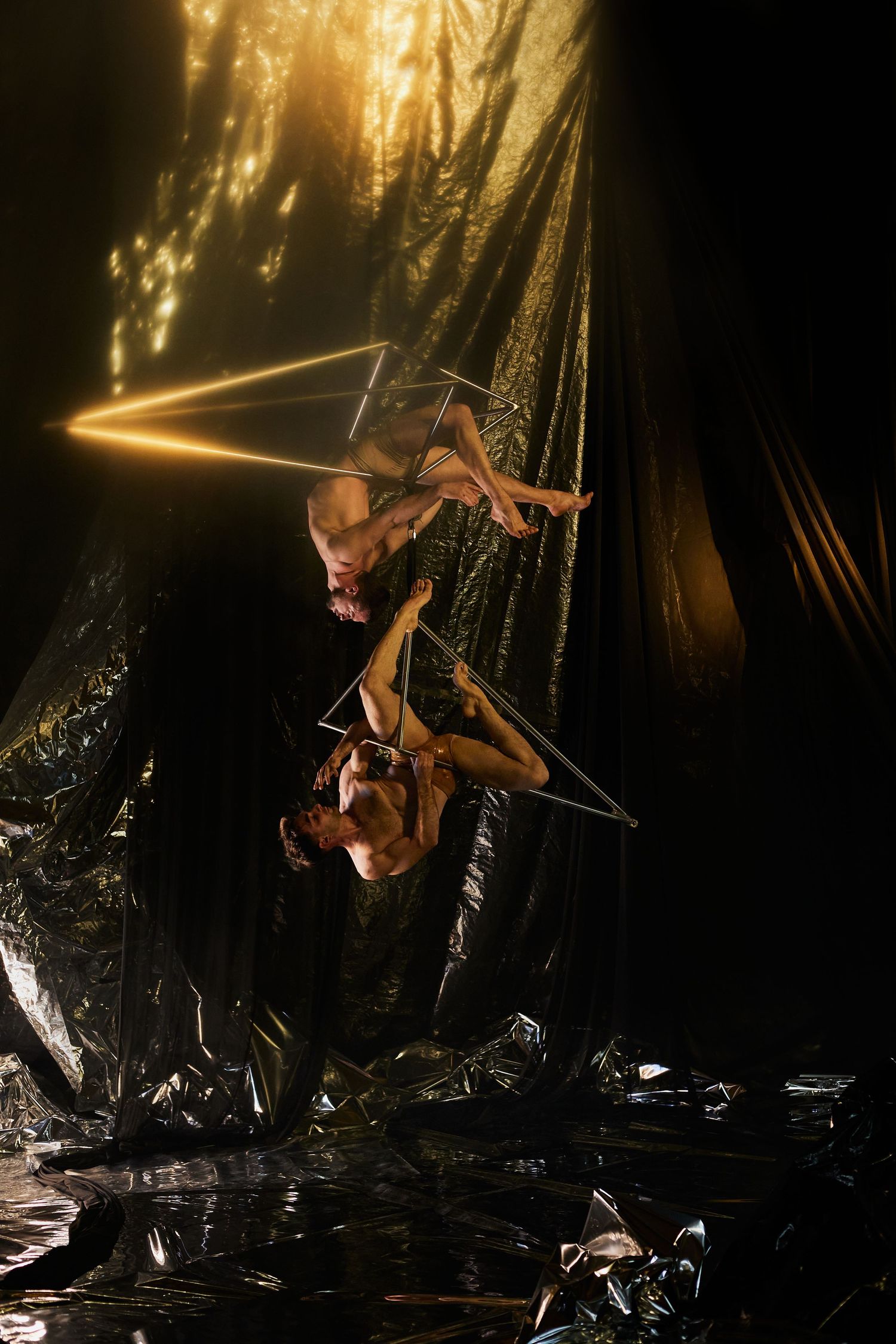 Acrobatic performer suspended on illuminated triangular apparatus against dark background with dramatic lighting effects.