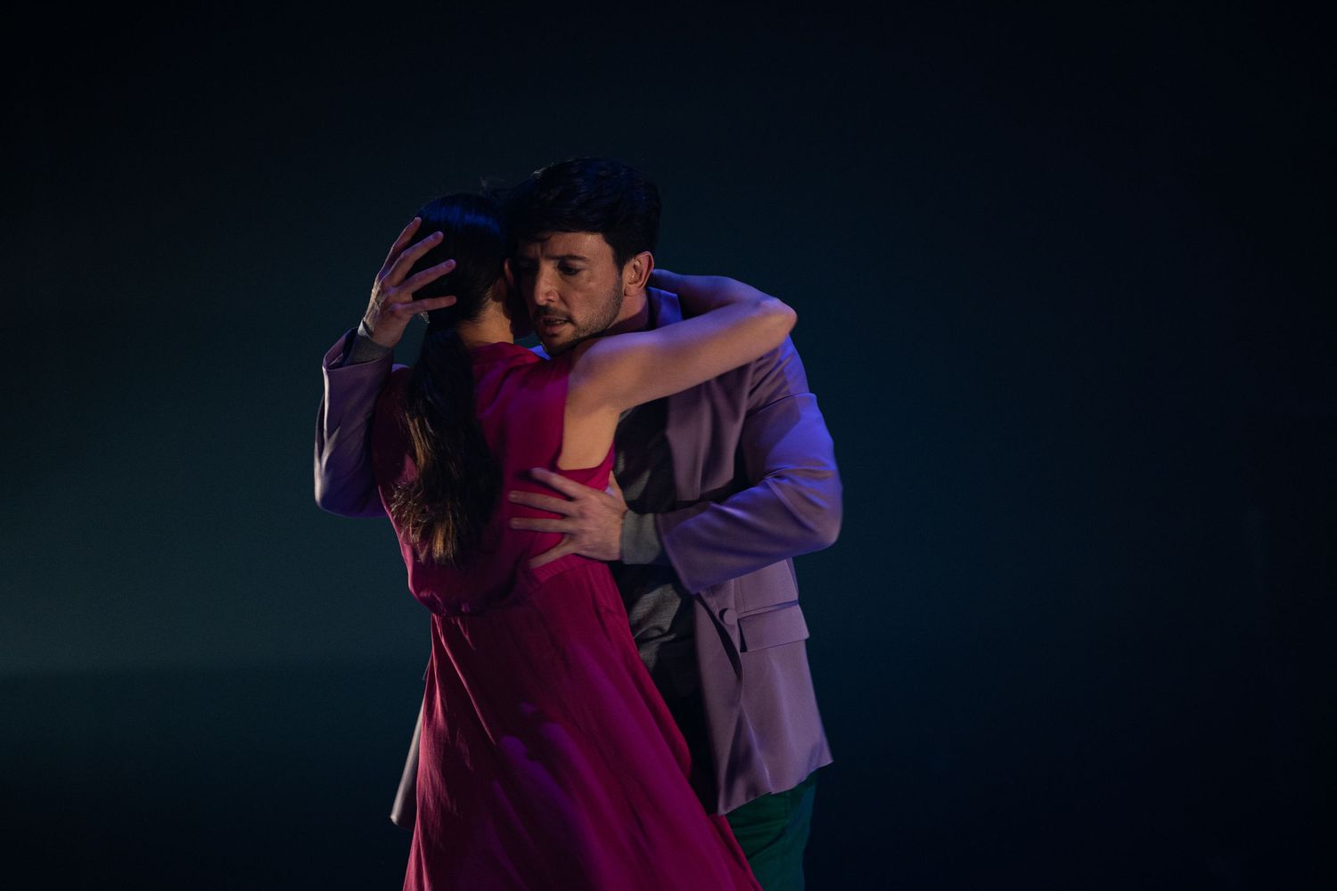 Two dancers perform an intimate partnered dance with dramatic purple and blue lighting on a dark stage.