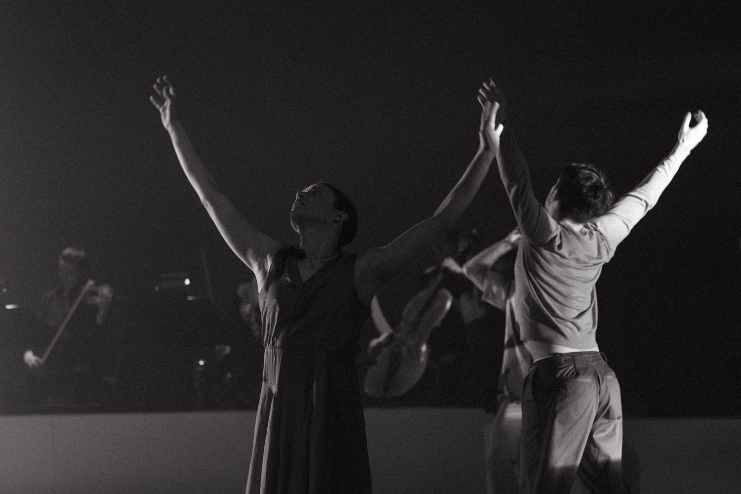 Silhouettes of dancers with raised arms performing on stage in black and white dramatic lighting.