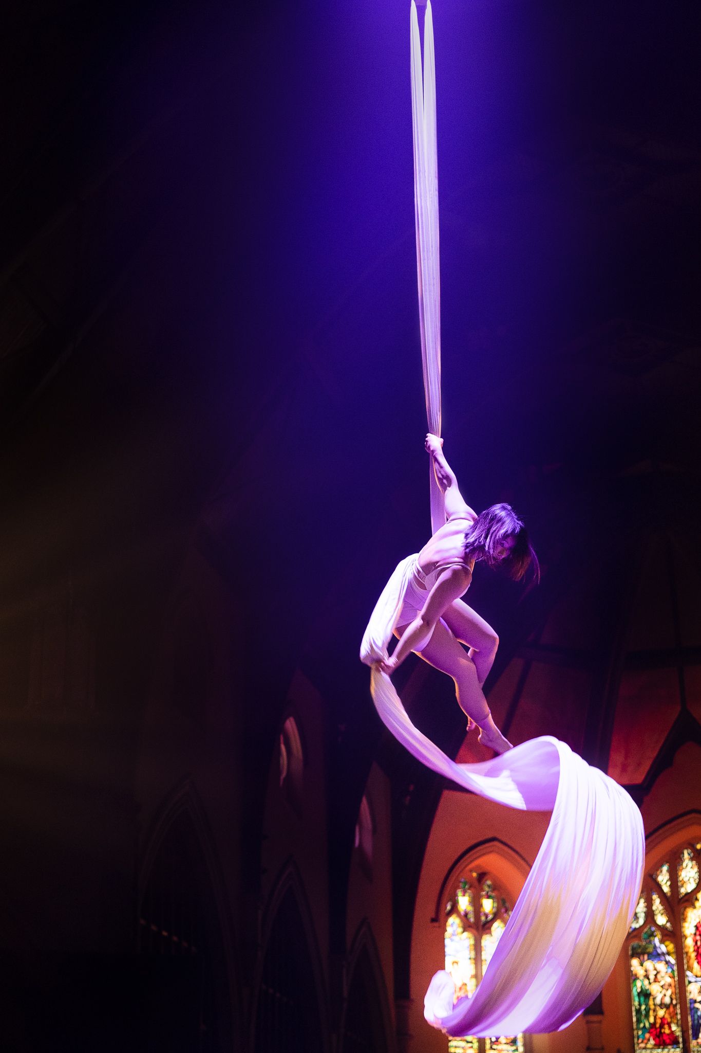 Aerial silk artist performs elegant moves with flowing white fabric against a deep purple-lit theatrical backdrop.