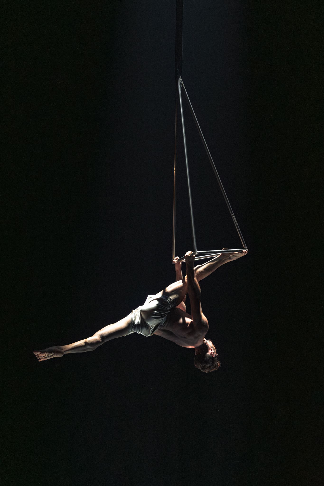 Aerial acrobat performs a dramatic split pose while suspended from thin ropes against a dark background.
