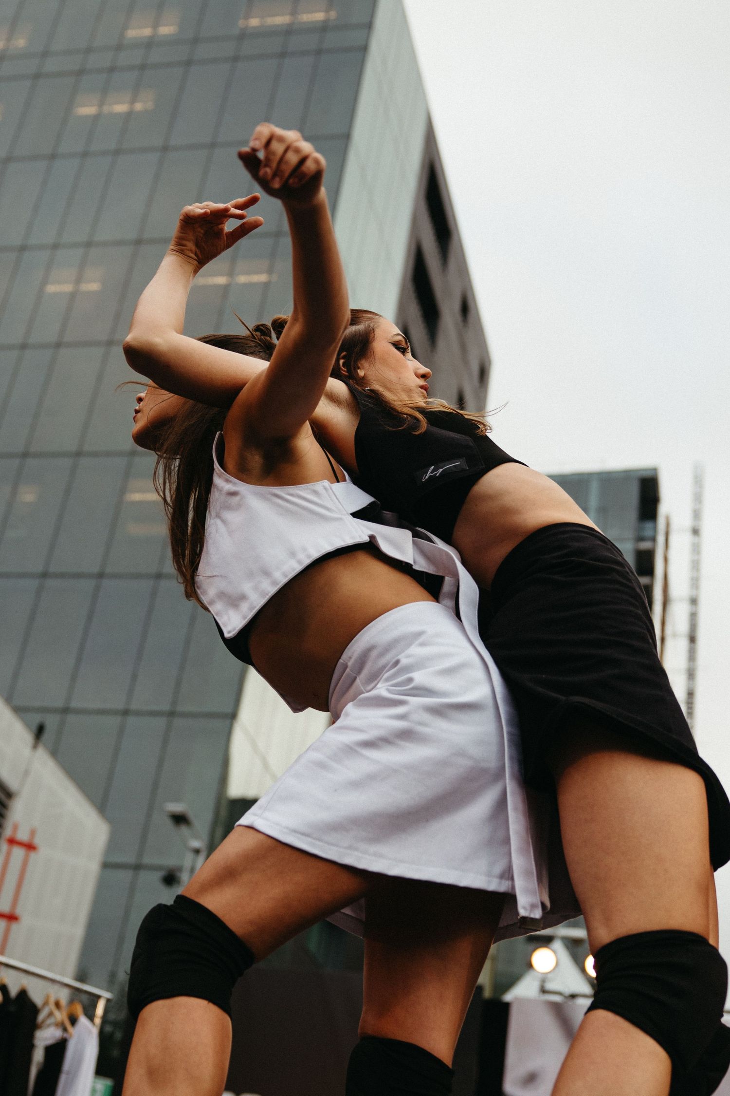 Two dancers in athletic wear performing dynamic dance moves against an urban backdrop with modern buildings.