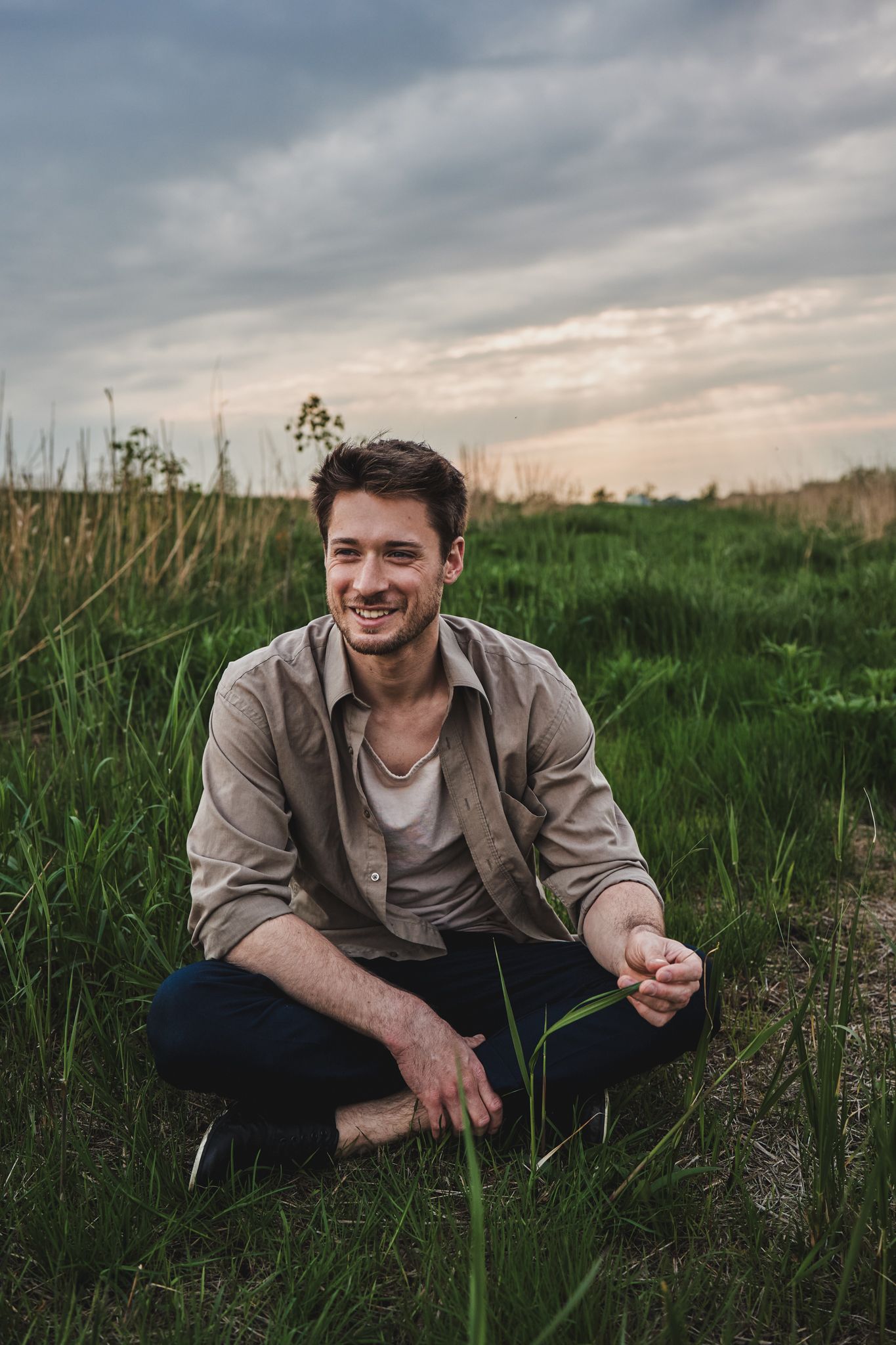 A person sits cross-legged in a grassy field at sunset wearing a beige button-down shirt and dark pants.