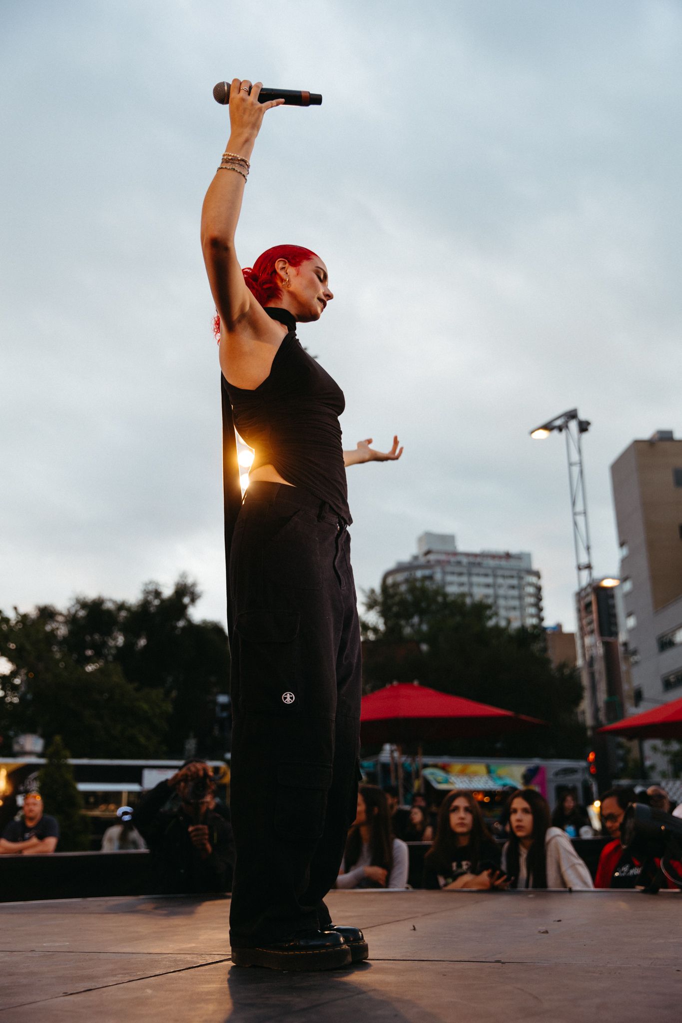 A performer in black clothing holds a microphone up while standing on an outdoor stage at dusk with city buildings in background.