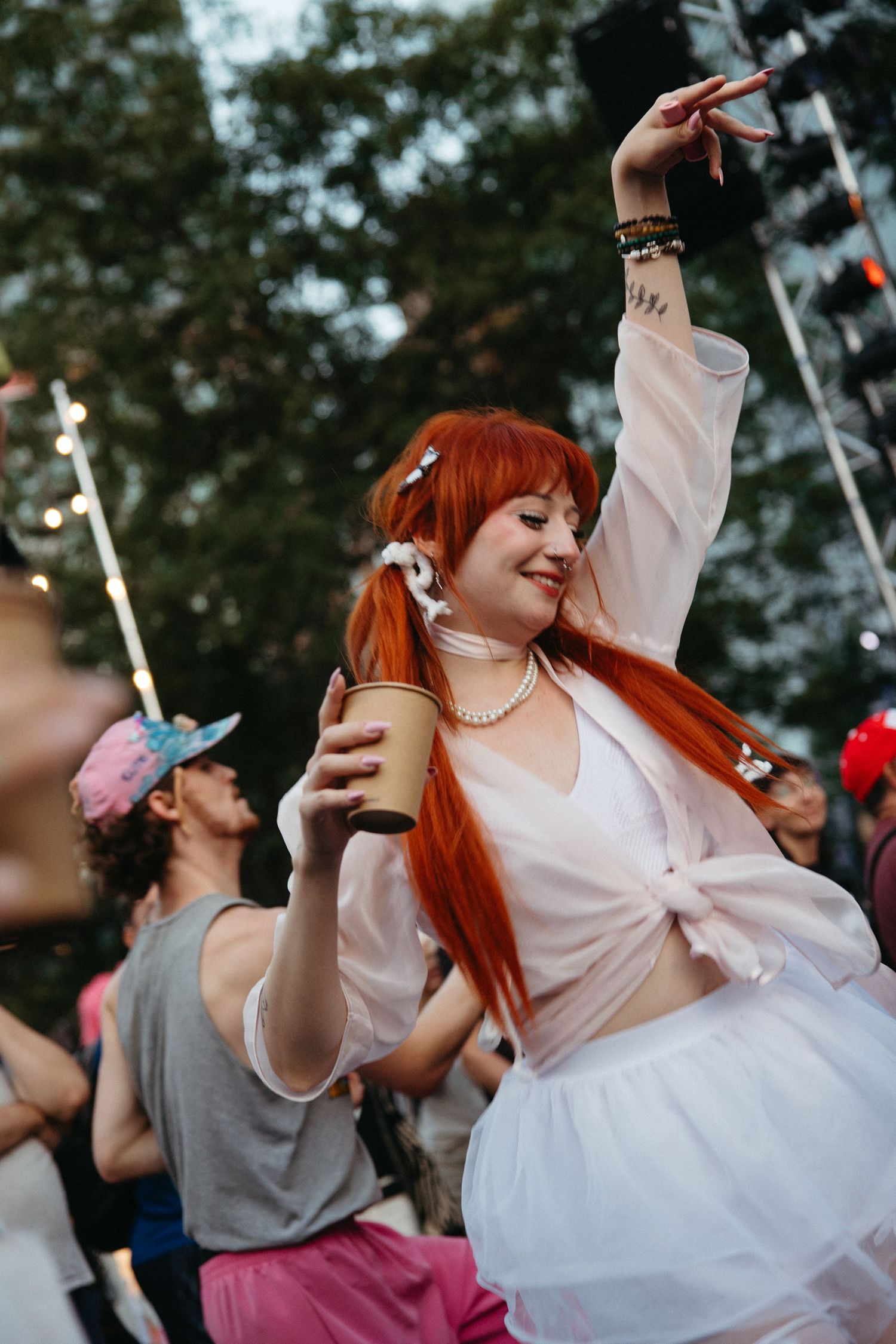 A festival goer with bright red hair dances joyfully while holding a drink at an outdoor summer event.