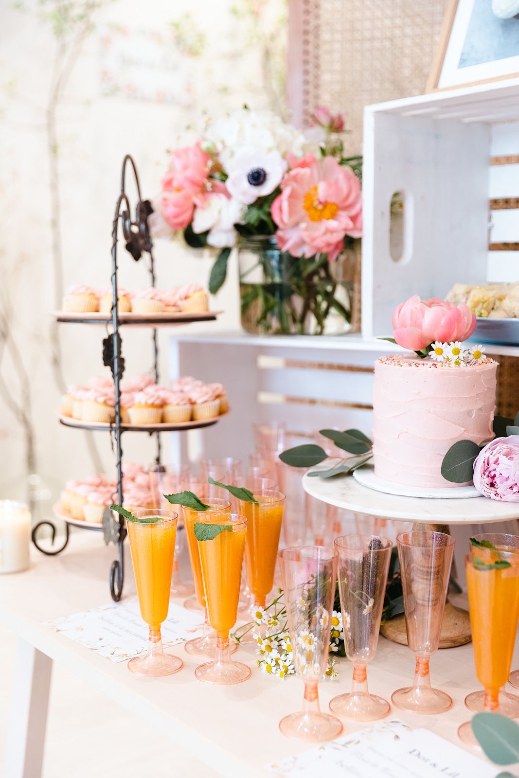 Elegant dessert display with pink cake, champagne flutes filled with orange cocktails, and fresh flowers in soft lighting.