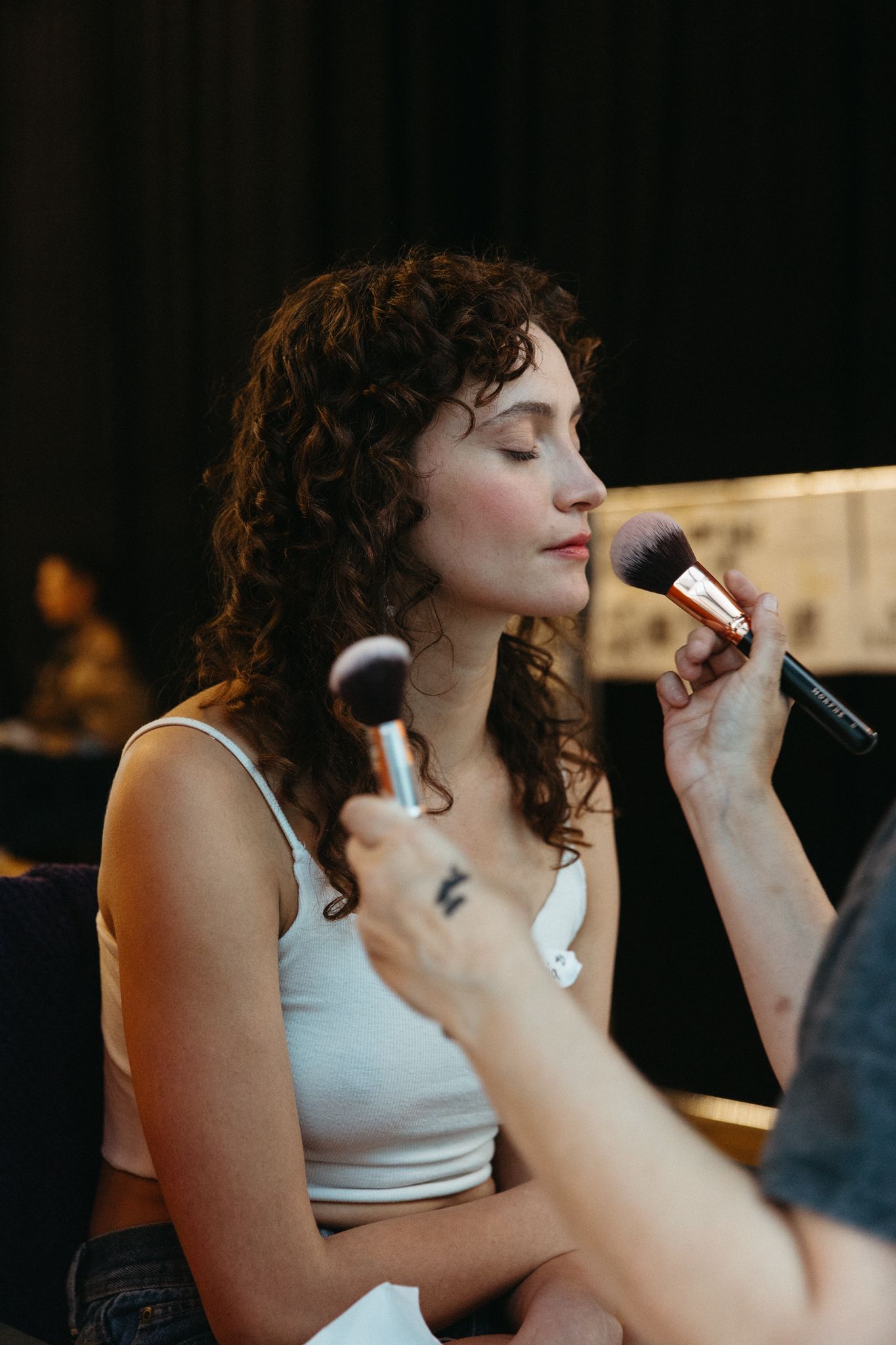Makeup artist applies cosmetic product with professional brushes during beauty session in dimly lit setting.