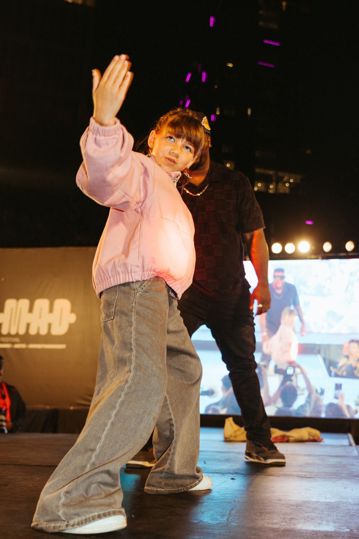 A performer in a pink sweater and baggy jeans strikes a dynamic pose on stage with stage lights and screens in background.