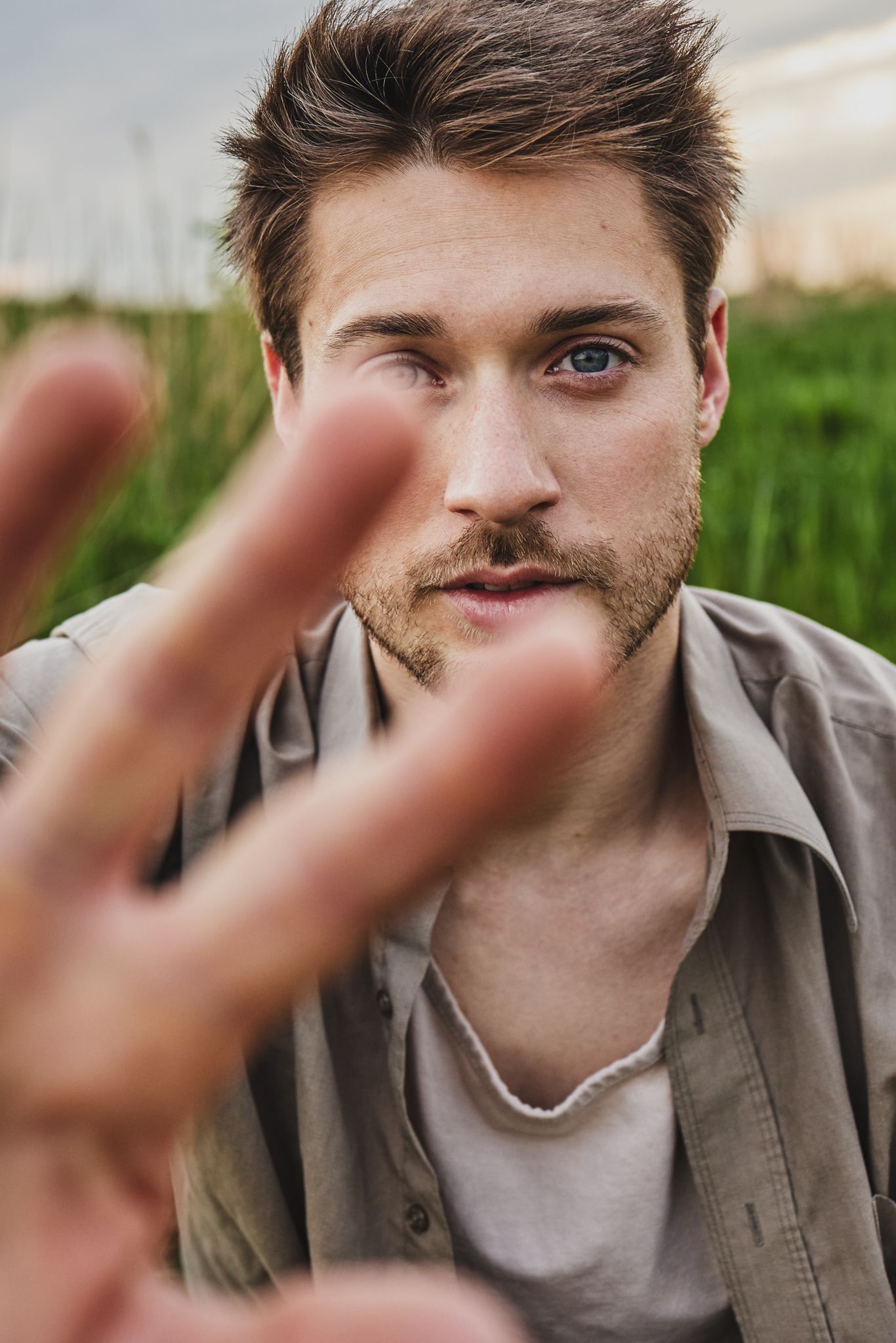 Close-up portrait with a hand reaching toward camera against green field background in natural lighting.