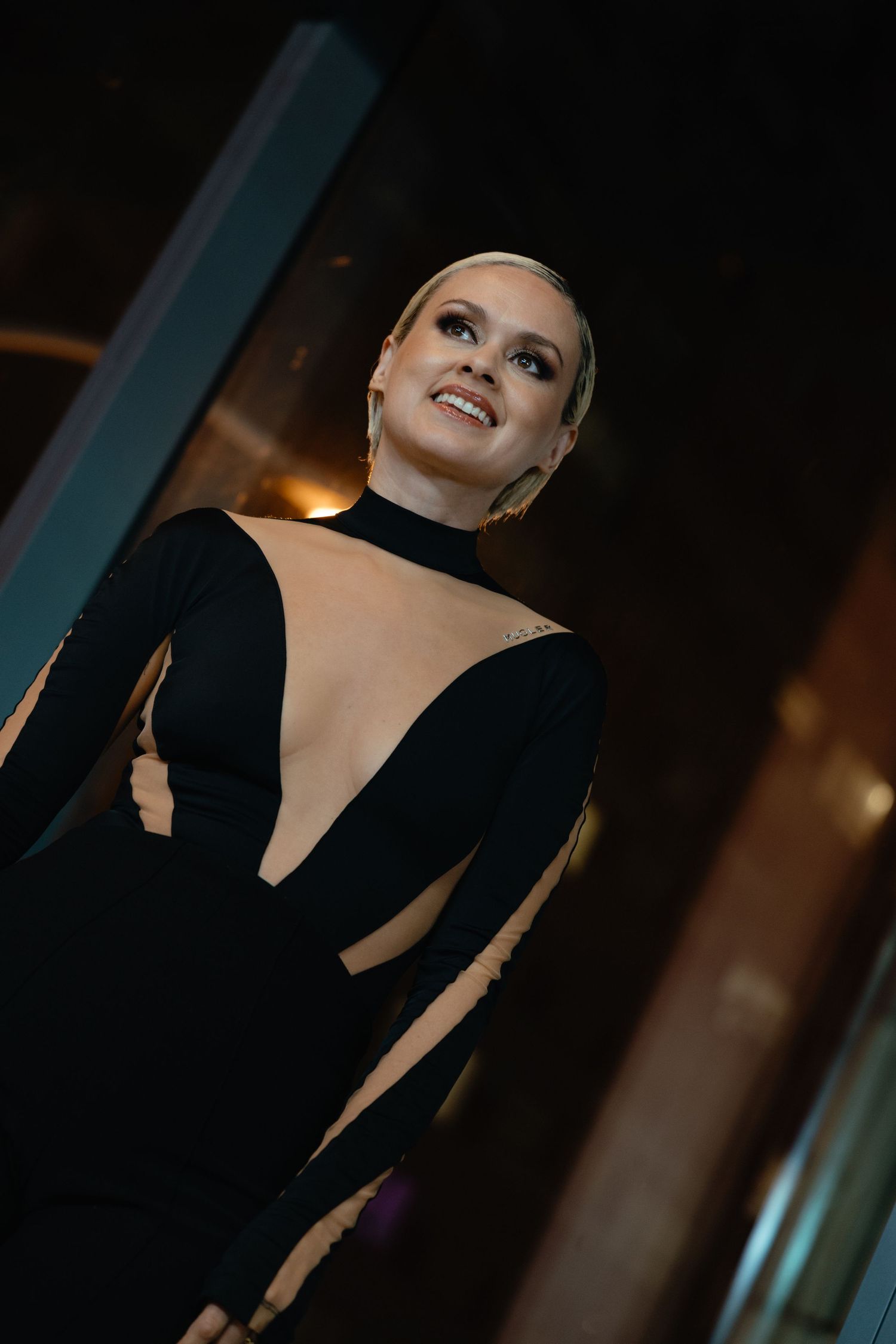 Elegant figure in a dramatic black evening gown with strategic cutouts poses against dark backdrop with soft lighting.