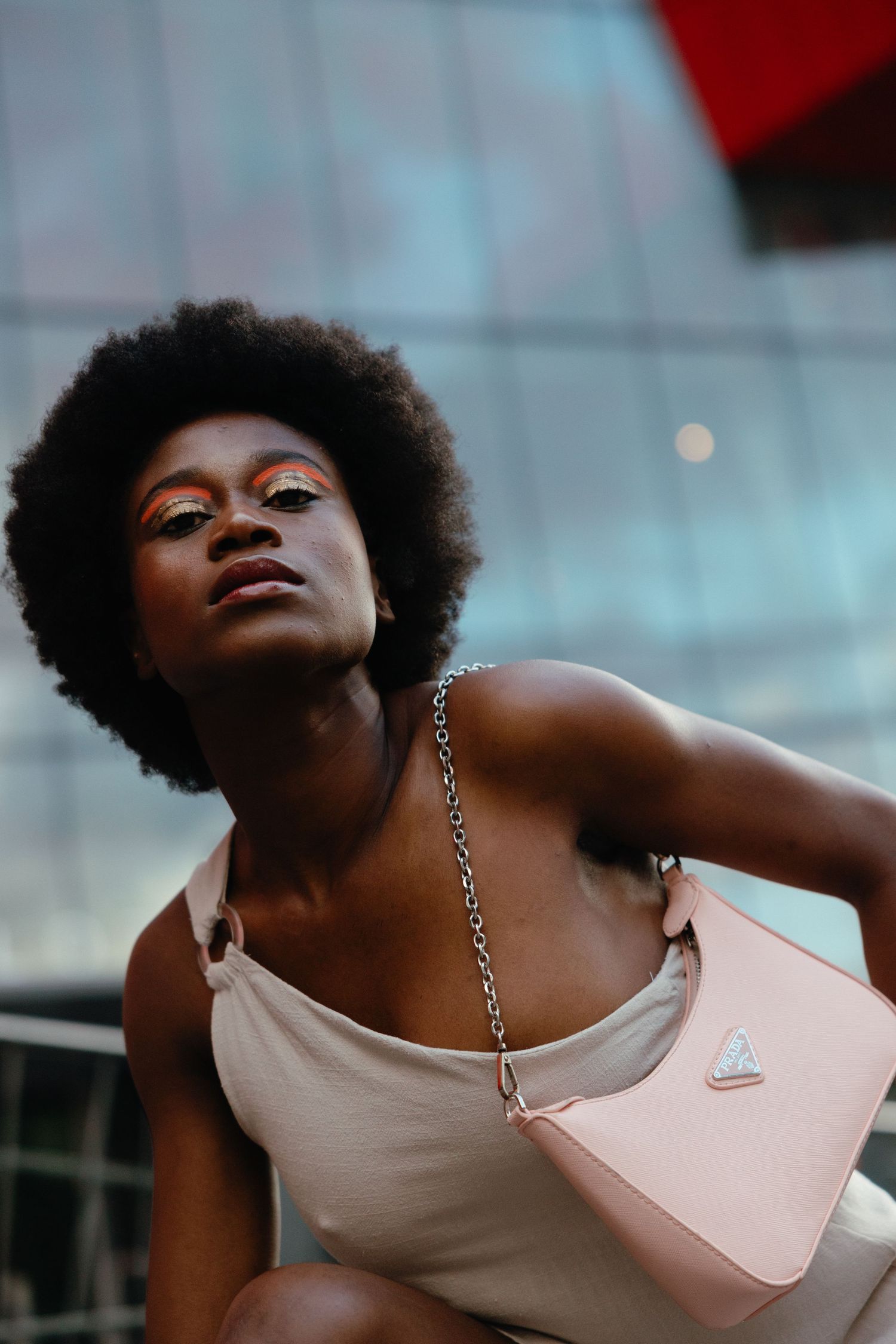 Portrait of a person with natural Afro hair wearing a light top and pink handbag against an urban background.
