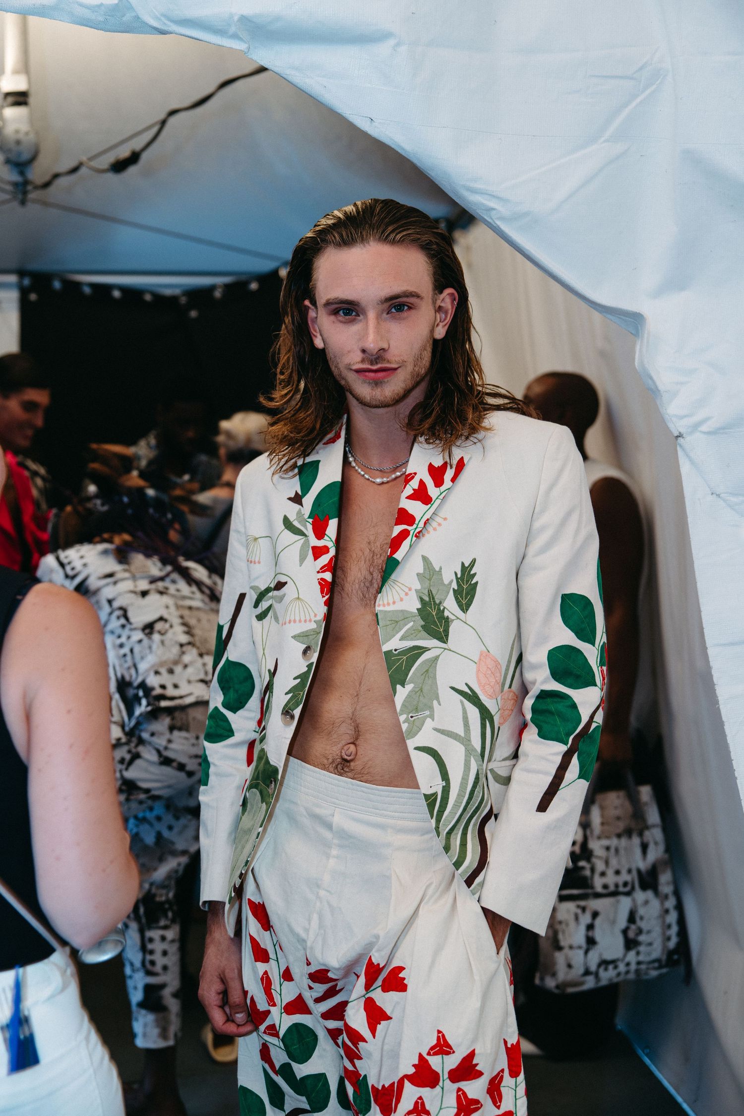 A model wears a white botanical print suit with green and red floral designs backstage at a fashion show.