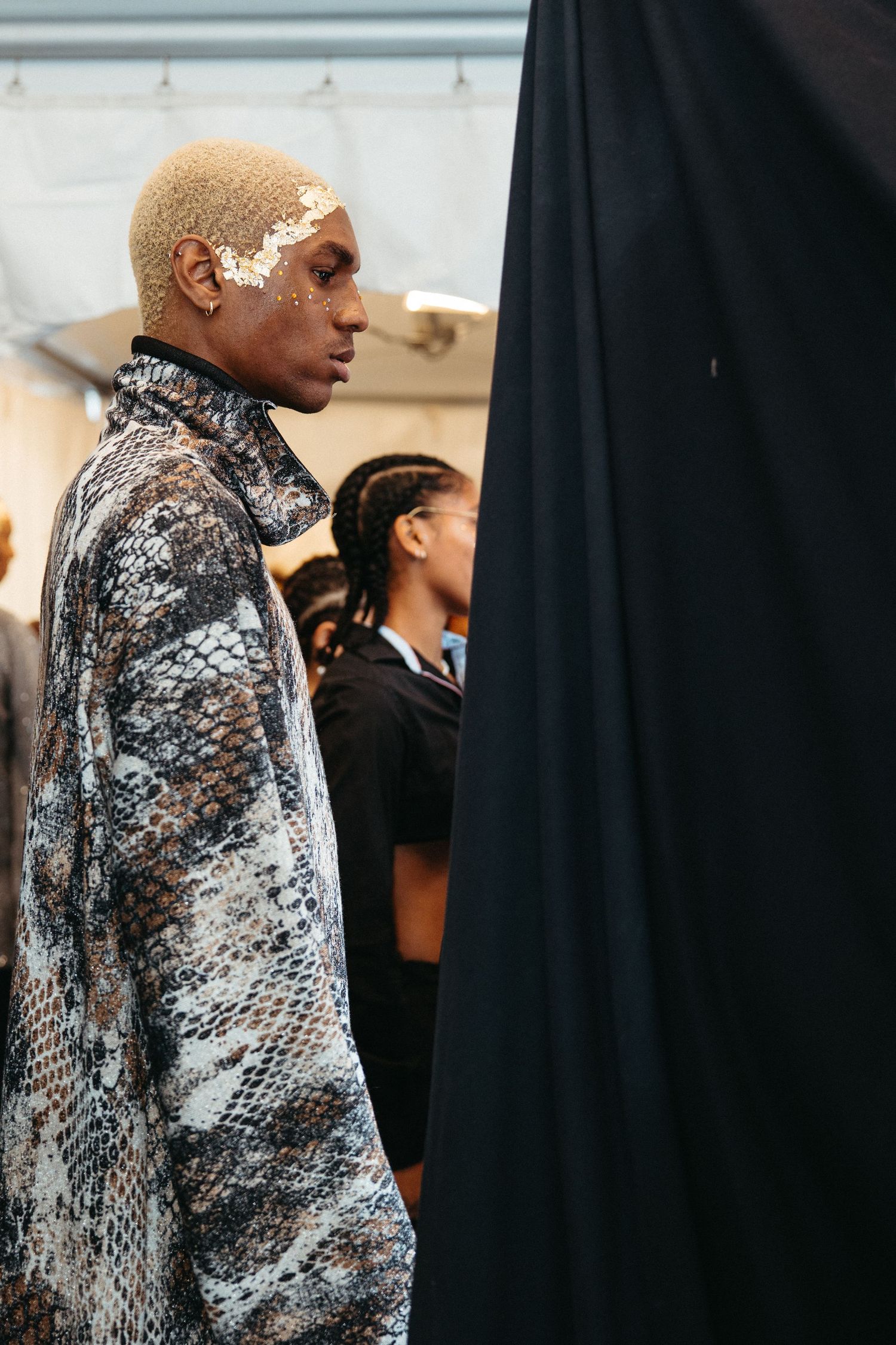 A person wearing a shimmering silver snakeskin print jacket stands against a black backdrop at a formal event.