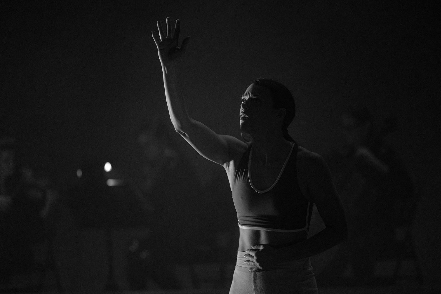 Black and white image of a performer in athletic wear with raised hand performing on a darkened stage.