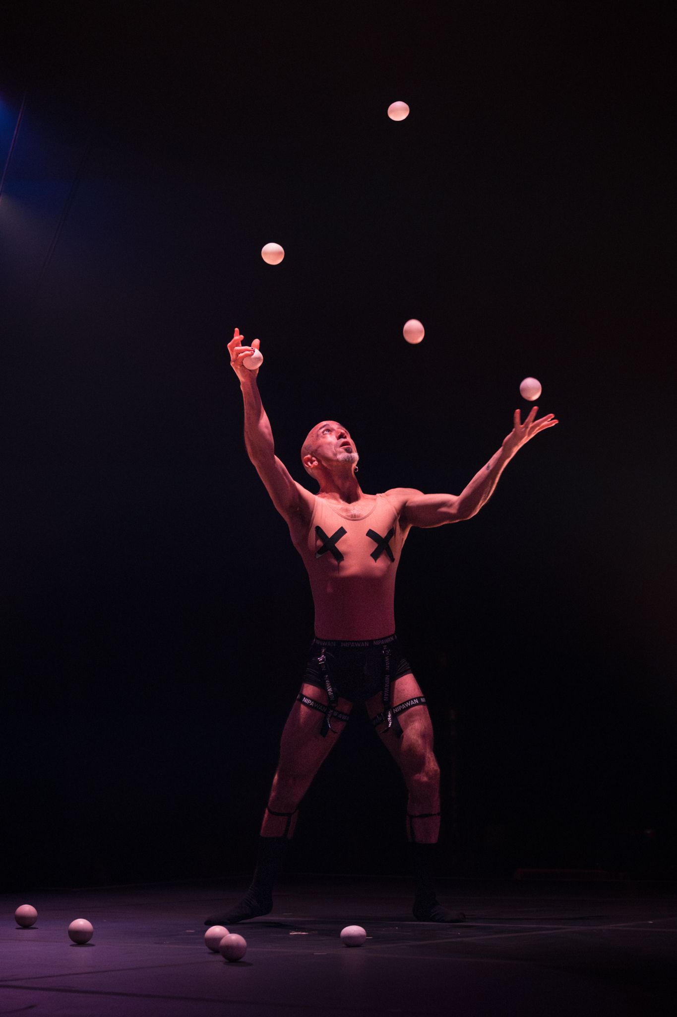 Performer in dark setting tosses juggling balls upward while making dynamic movements on stage.