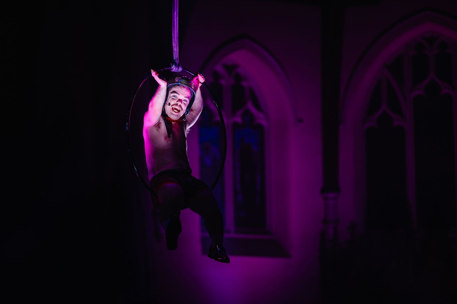 A performer illuminated by purple lights hangs from aerial silks against a dark background with gothic arches.