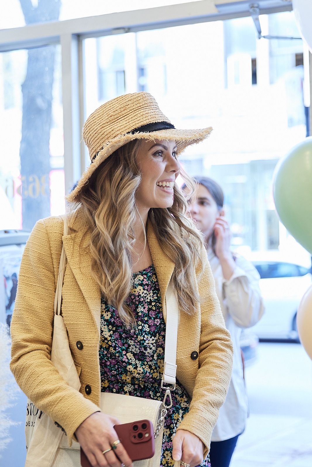 Woman in straw hat and yellow blazer wearing floral dress smiles while standing in bright indoor space.