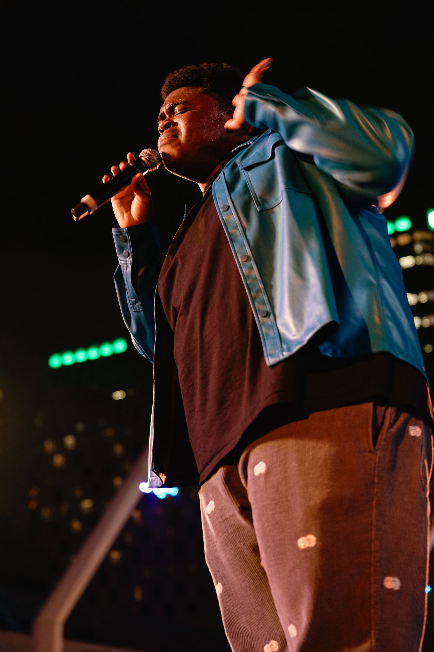 A performer in a brown and light blue outfit sings passionately on stage with dramatic lighting in the background.
