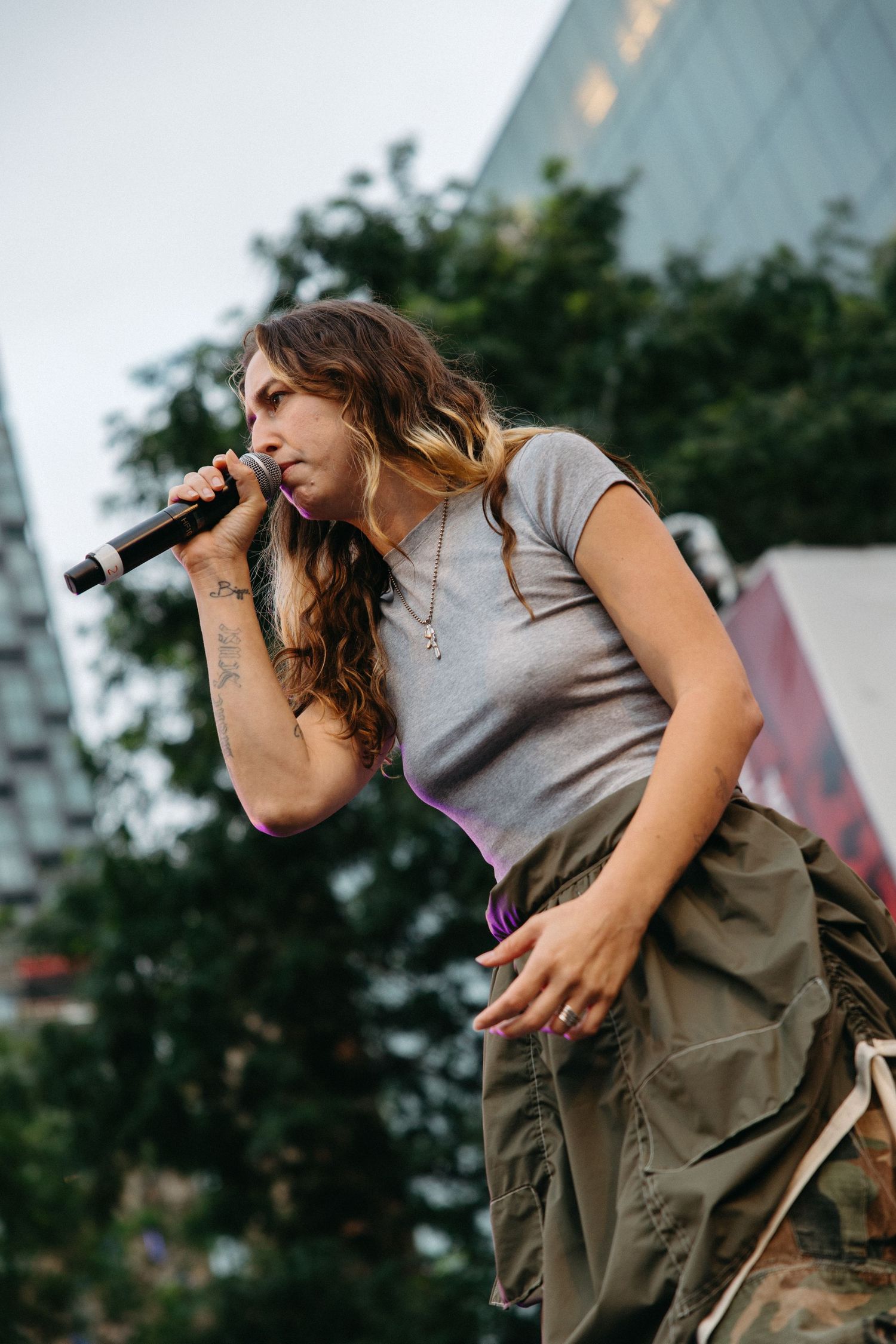 A performer in a gray t-shirt and cargo pants sings passionately into a microphone at an outdoor concert venue.