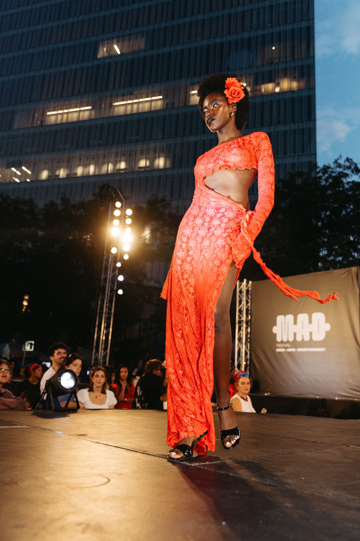Model showcases stunning coral lace two-piece ensemble on outdoor runway with urban skyline backdrop.