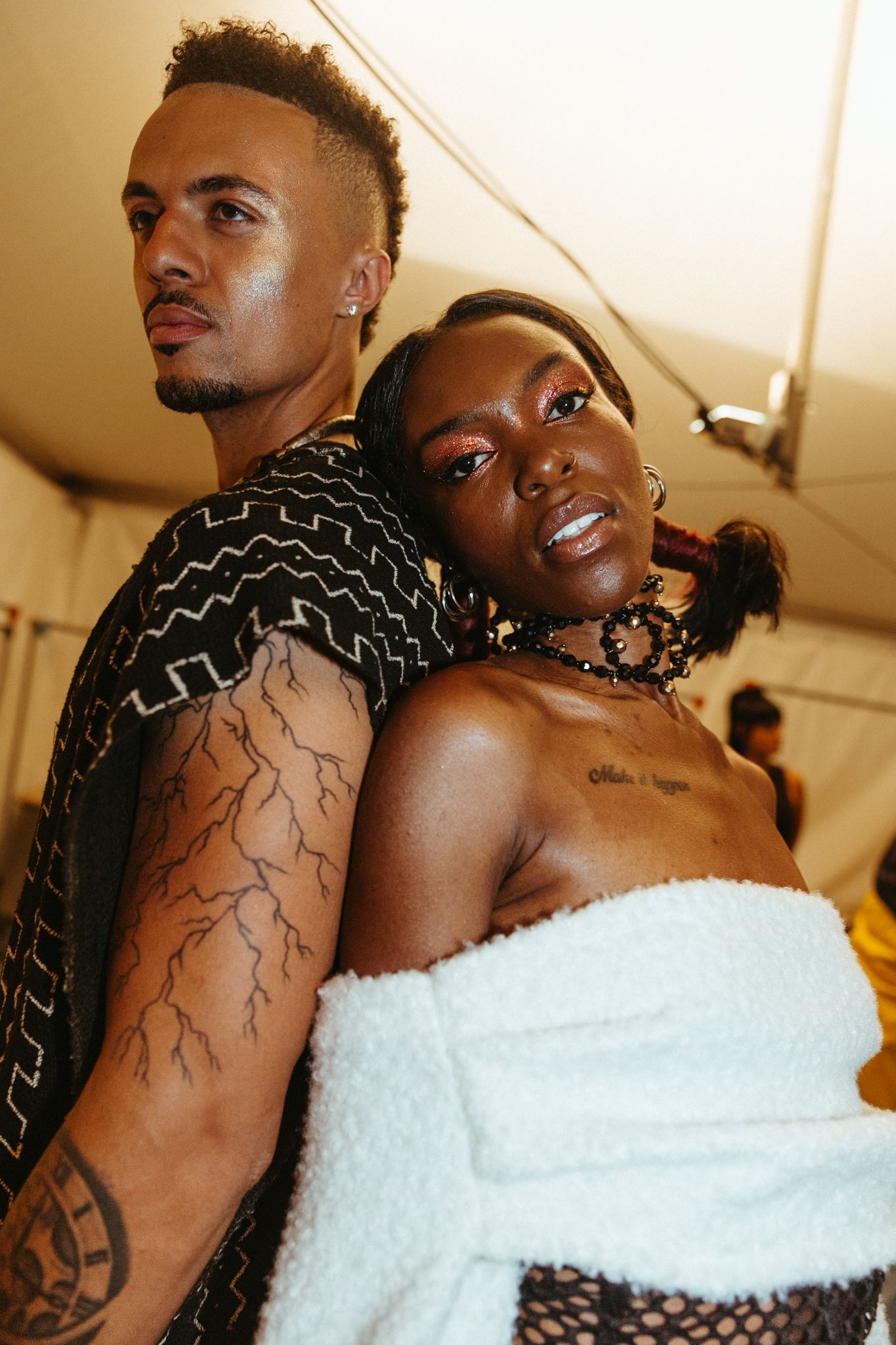 Two performers pose together backstage, one wearing a patterned top with tattoos and one in a white off-shoulder outfit.