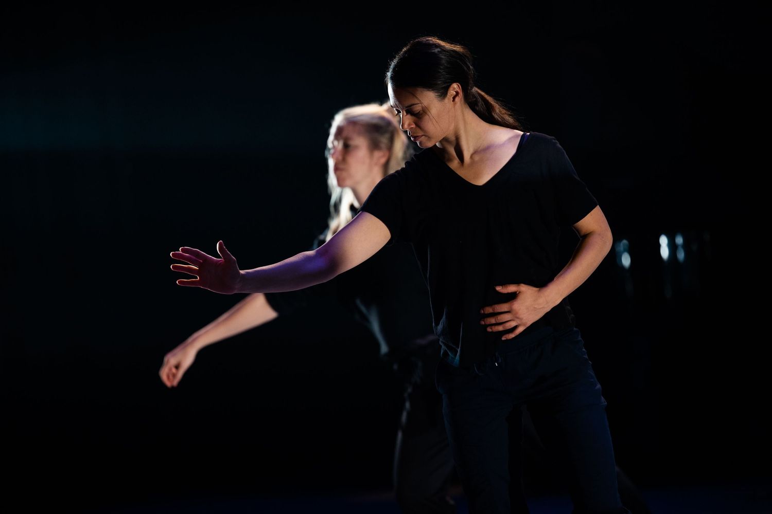 Two performers rehearse a dramatic dance lift sequence on a dimly lit stage.