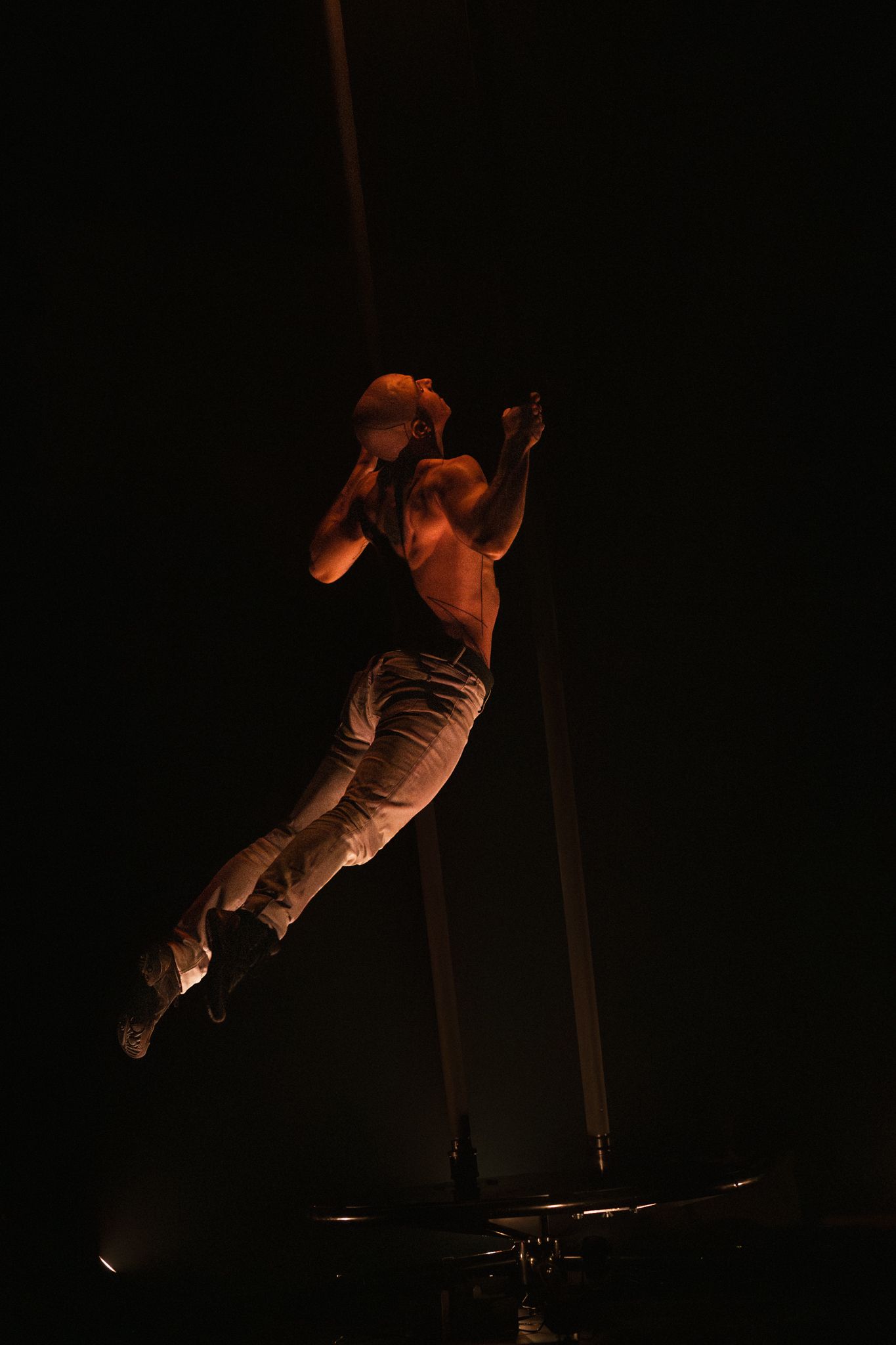 Aerial performer executes dramatic pose while suspended on poles under warm amber lighting.