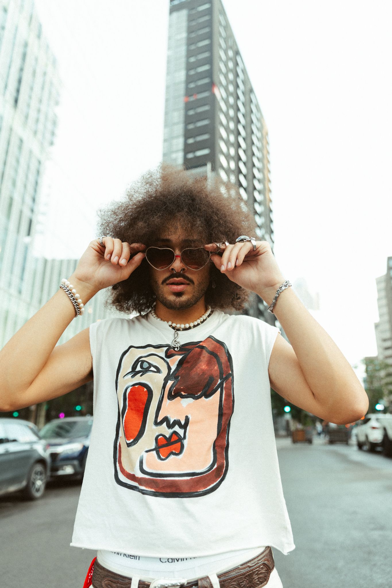 Person with curly hair wearing sunglasses and white graphic t-shirt posing against urban cityscape backdrop.