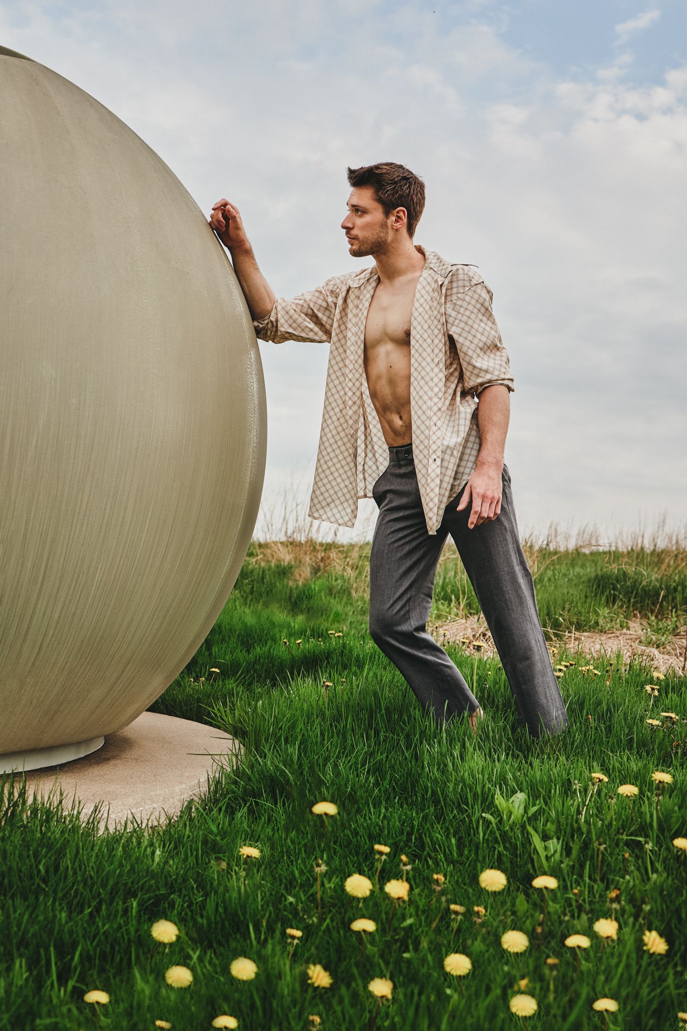 A person in an open shirt poses against a large sphere sculpture in a grassy field with yellow wildflowers.