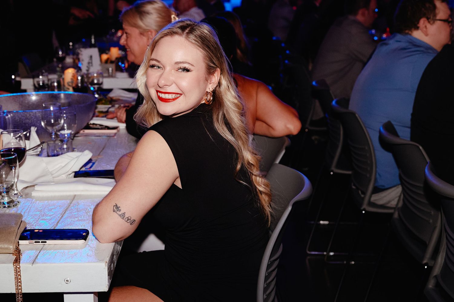 Guest in sleeveless black dress smiles at dinner party with table settings and dark ambient lighting in background.