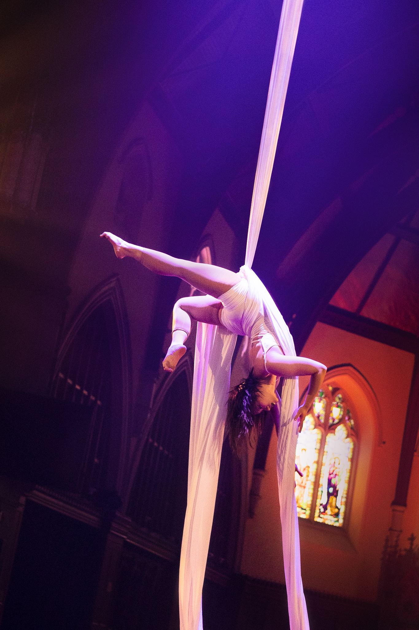Aerial performer in white fabric gracefully poses during a dramatic aerial silk performance in a dimly lit venue.